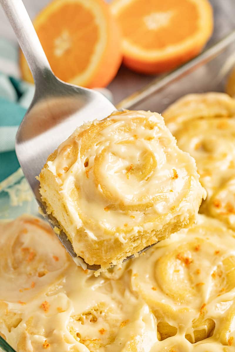 Orange Roll being held up by a metal spatula above a pan full of orange rolls.