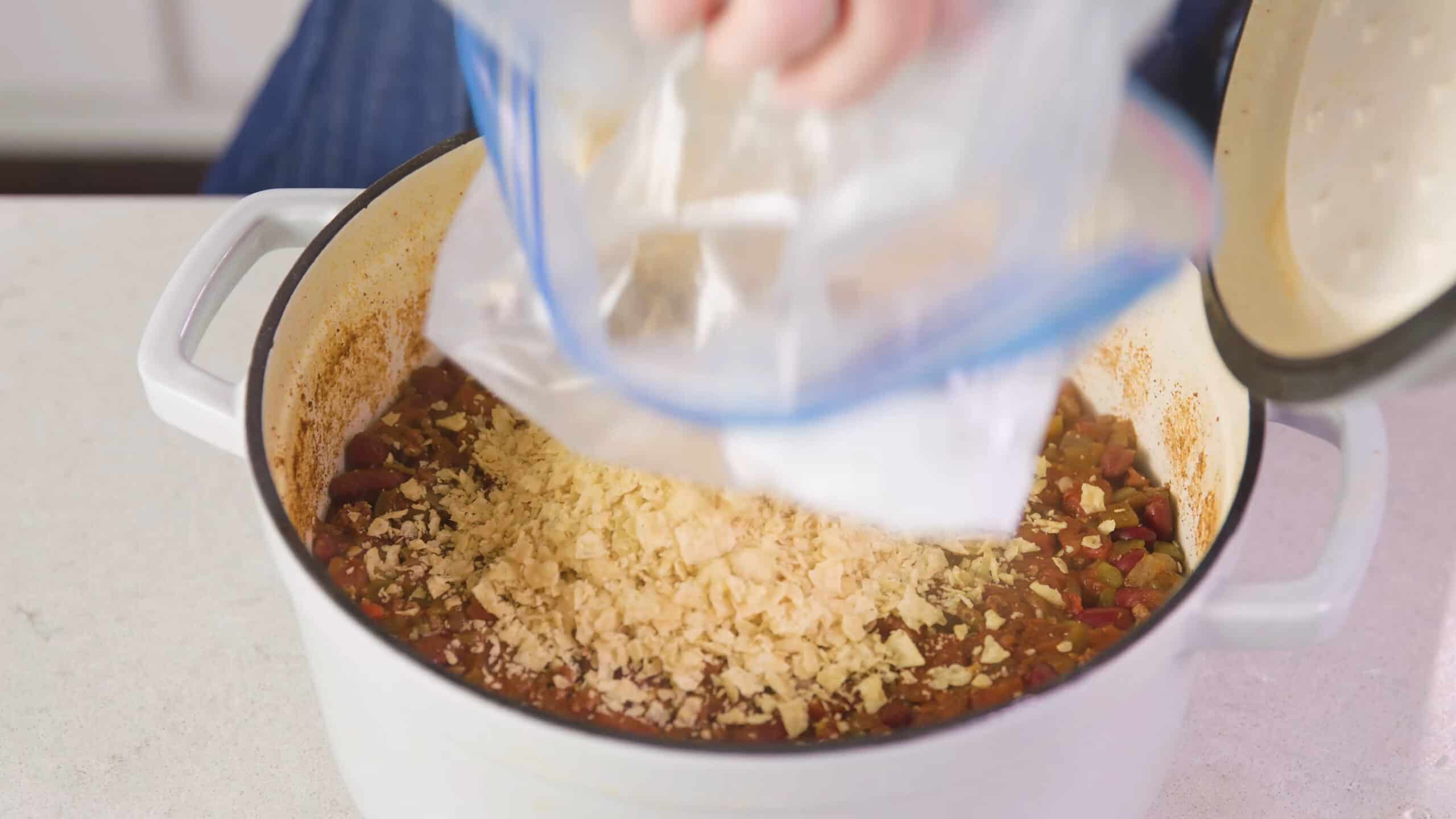 Angled view of white enamel cast iron pot filled with chili and crushed white corn tortilla chips added to ingredients from a large clear plastic bag, all on a marble countertop.
