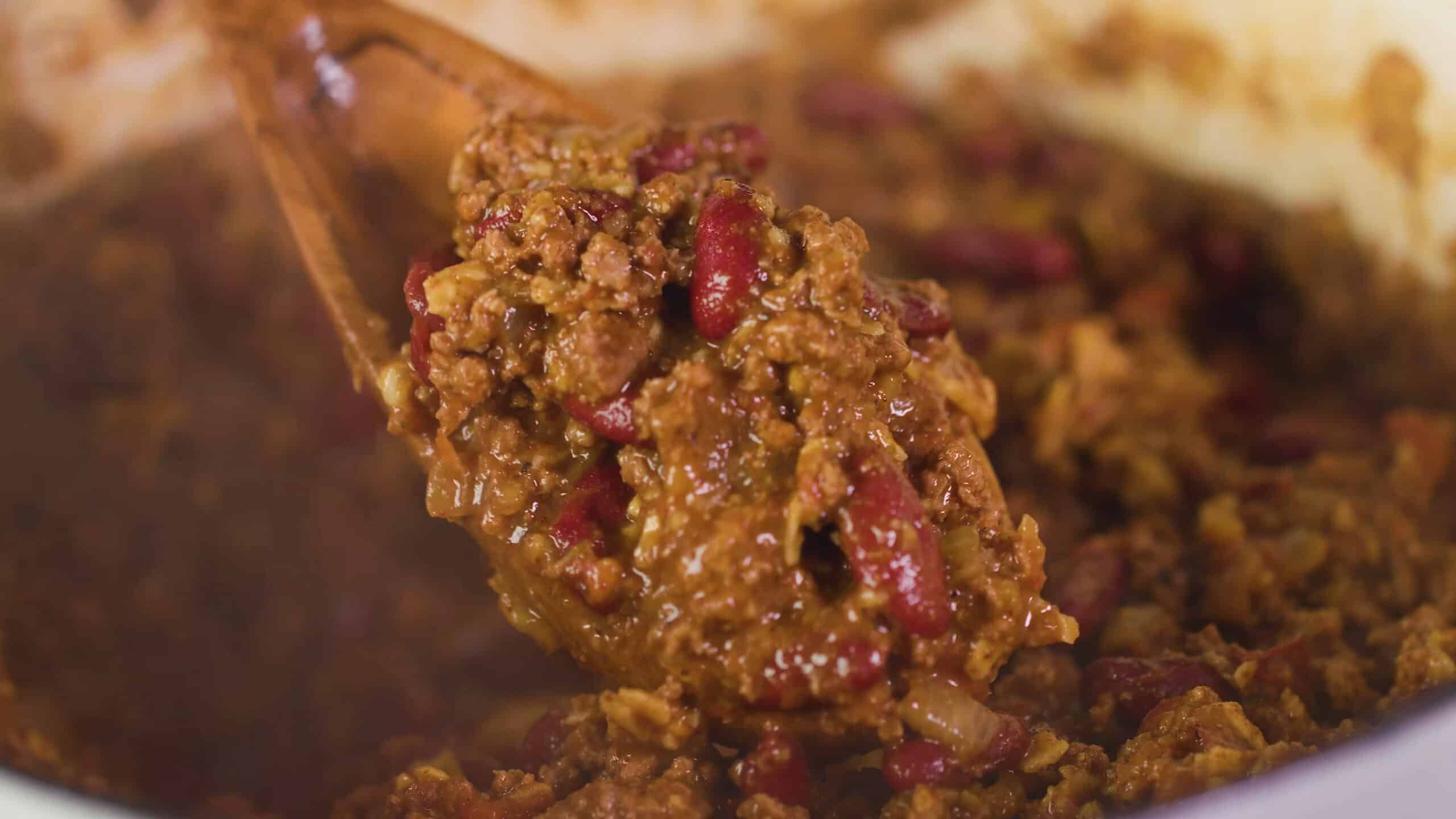 Close-up view of a large scoop of delicious chili on a wooden spoon with the rest of the chili making up the background.