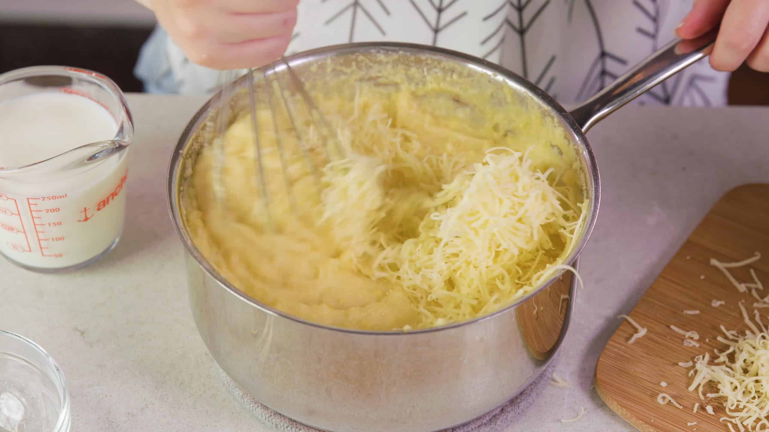 Angled view of a.silver pot on a countertop with white cheddar cheese being mix into the polenta ingredients with a whisk.