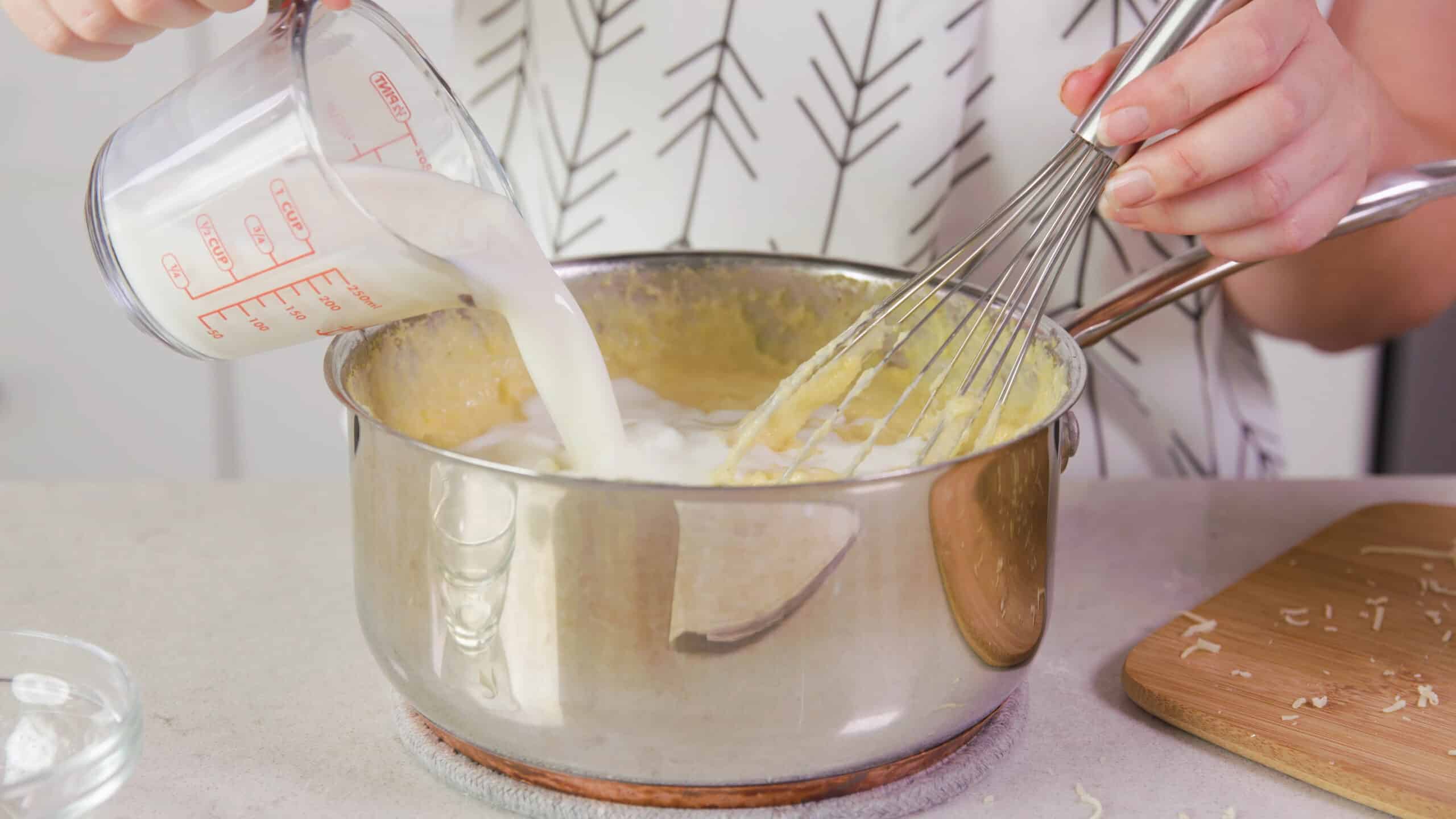 Angled view of silver pot on countertop filled with polenta mixture and pouring milk from a measuring cup and mixed with a wire whisk.