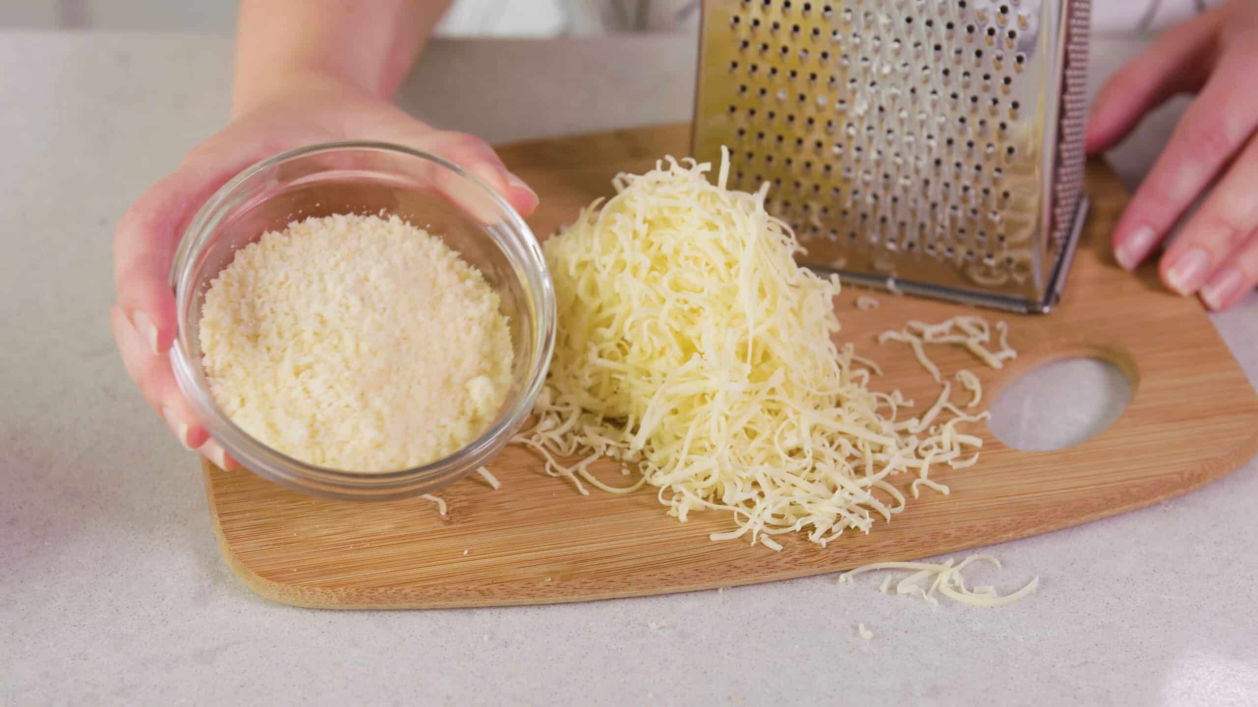 Using the small side of the cheese shredder, shred some white cheddar cheese onto a cutting board with a small clear bowl of parmesan cheese to the side.