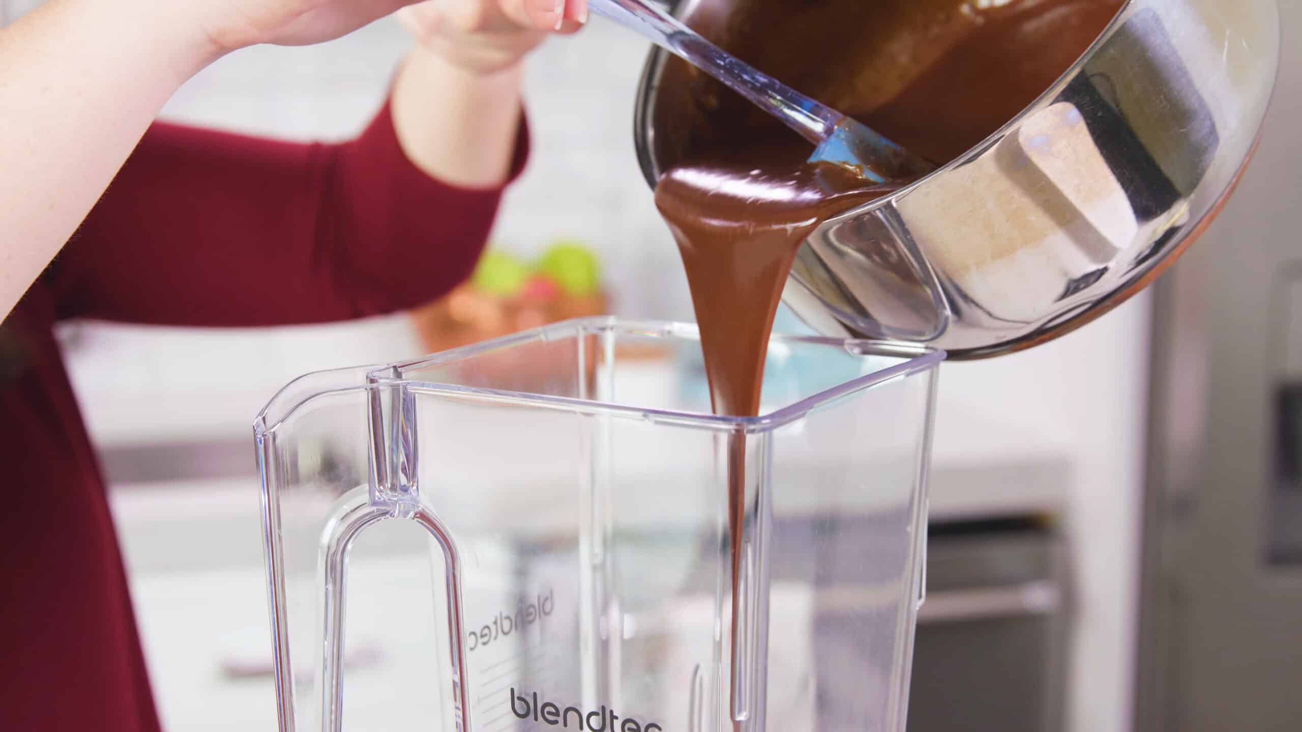 A silver saucepan pouring the chocolate sauce into a blender using a spatula.