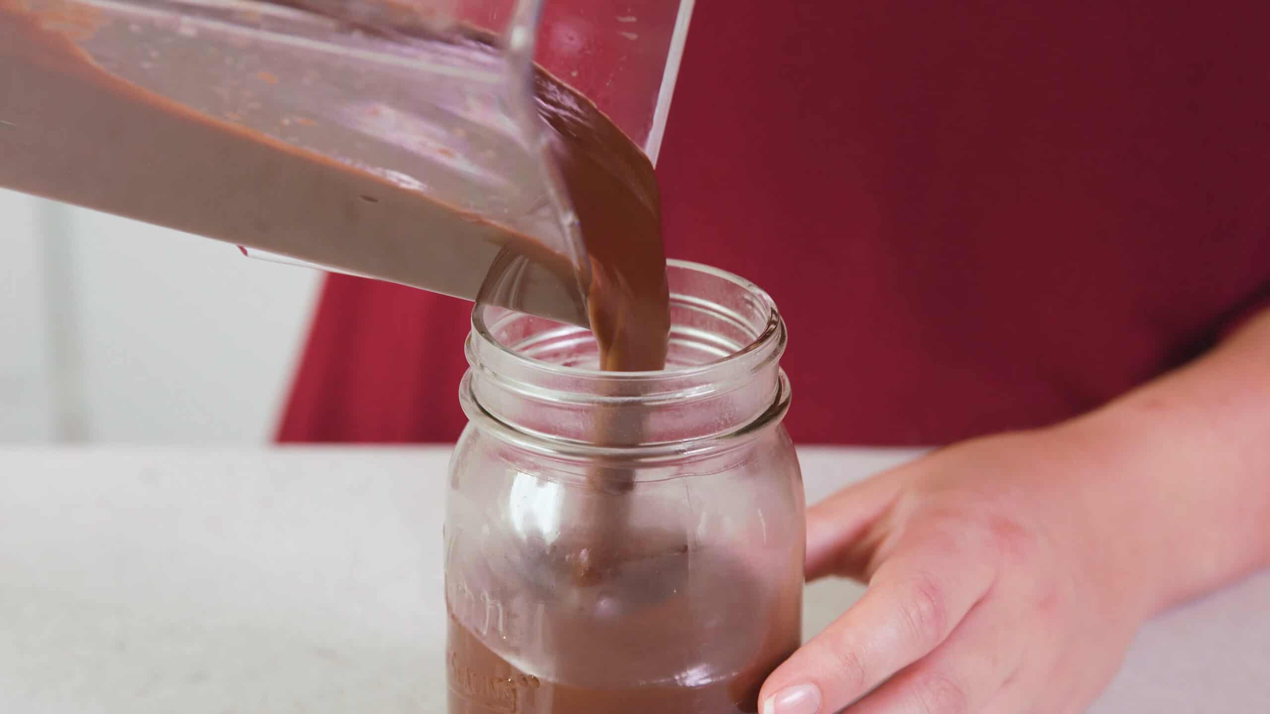 A blender pitcher pouring chocolate hot fudge sauce into a mason jar.