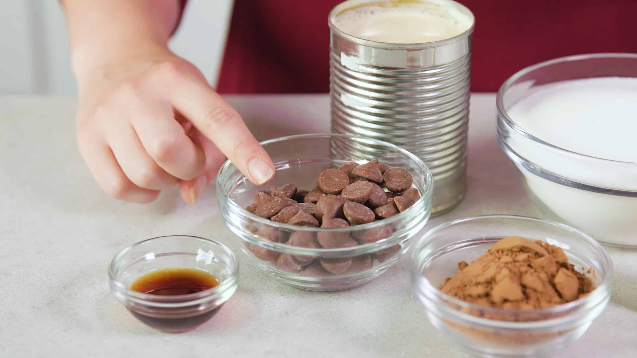 A series of ingredients laid out on a countertop including a clear dish of vanilla extract, cocoa powder in a clear dish, granulated sugar in a clear dish, a aluminum can of sweetened condensed milk. and some chocolate melting chips in a clear dish.