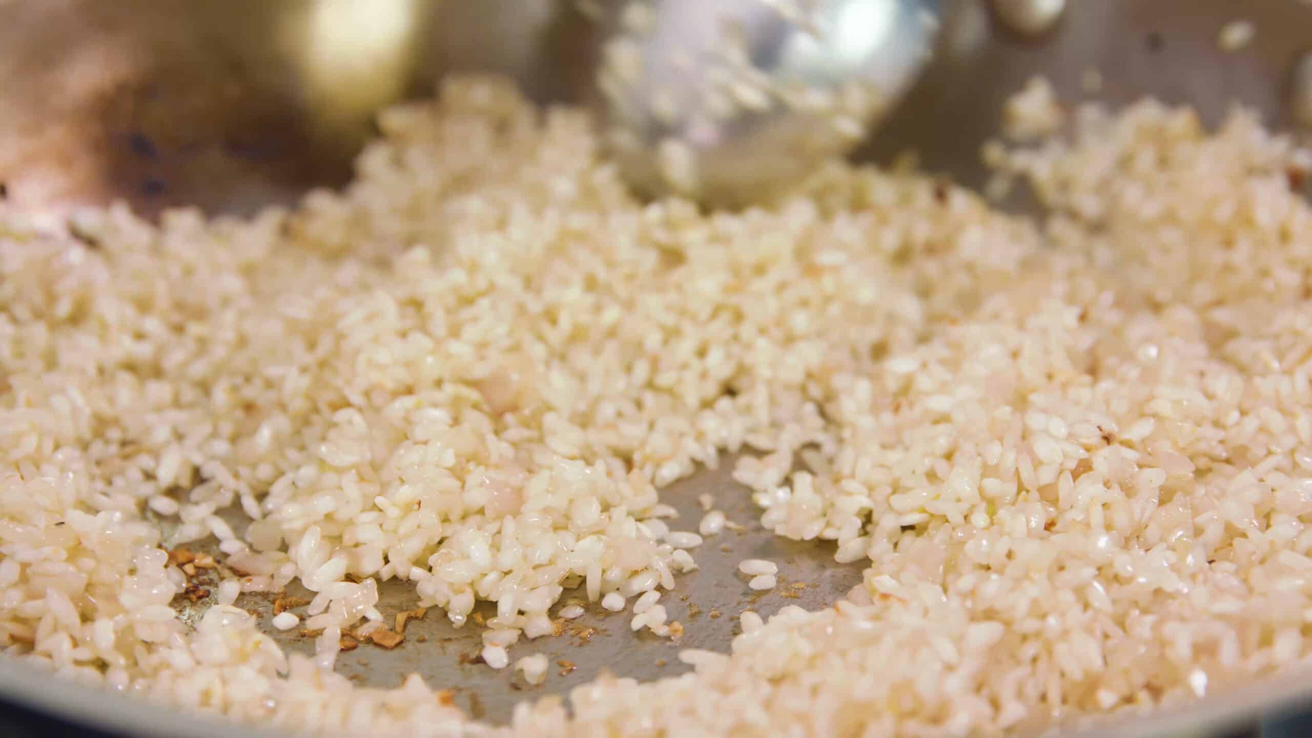Close-up view of risotto added to translucent minced onions in silver saucepan and mixed with silver spoon.