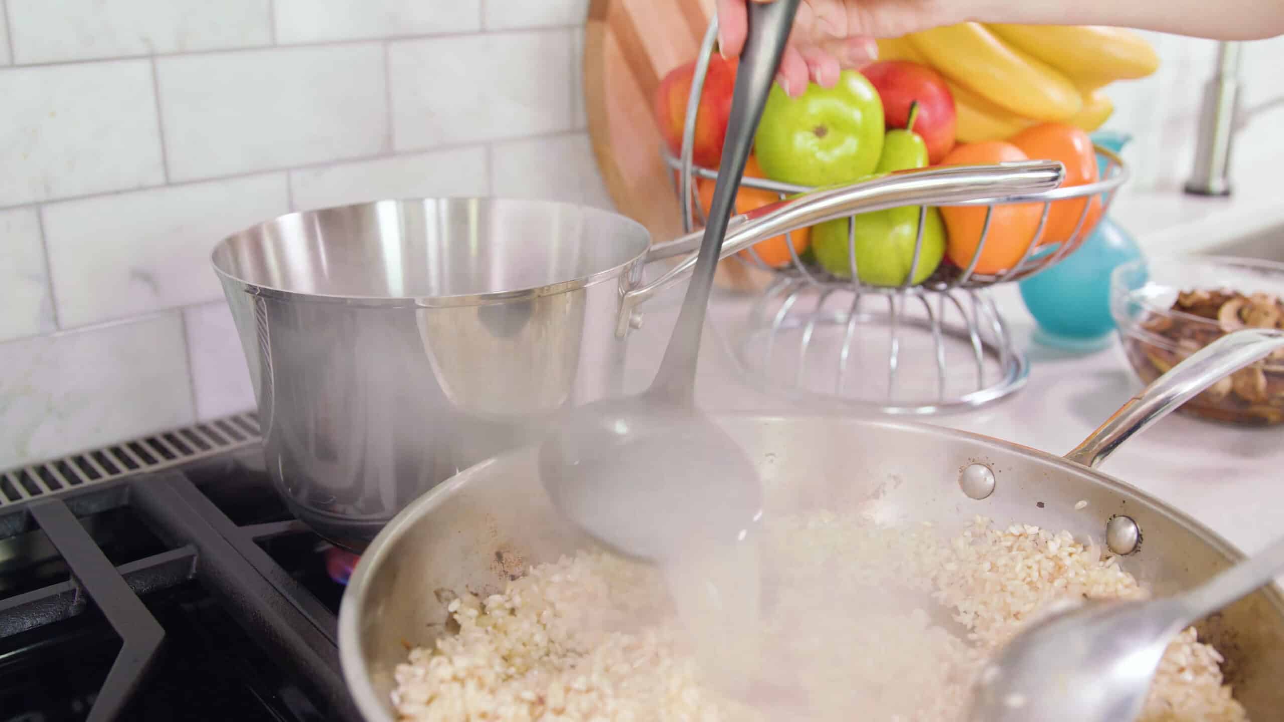 A silver saucepan on a stovetop with risotto and a ladle adding chicken stock to the saucepan from a silver pot.