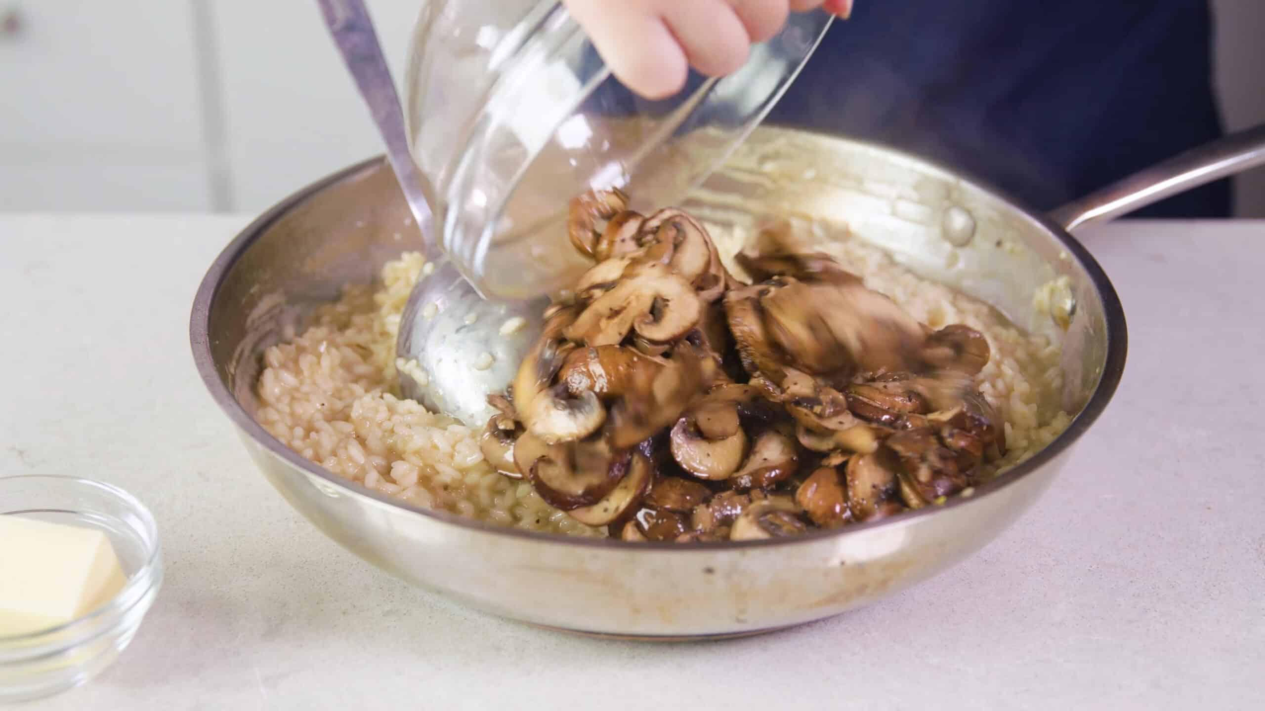 Silver saucepan on a countertop filled with cooked risotto and a large mixing bowl pouring sautéed mushrooms into the risotto mixture.