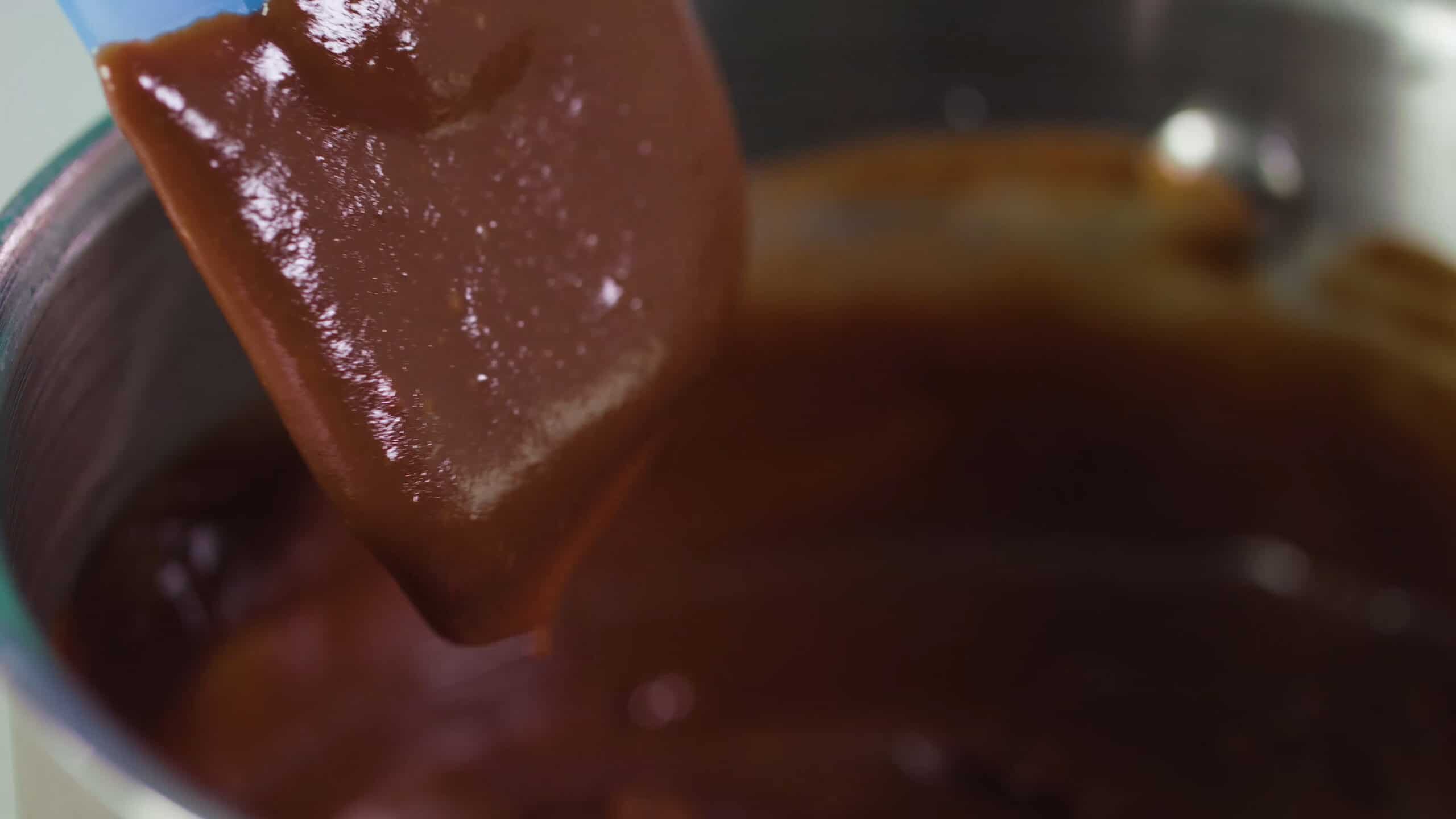 Close up view of plastic spatula covered in chocolate pudding to show texture with remaining pudding in silver pot in background.