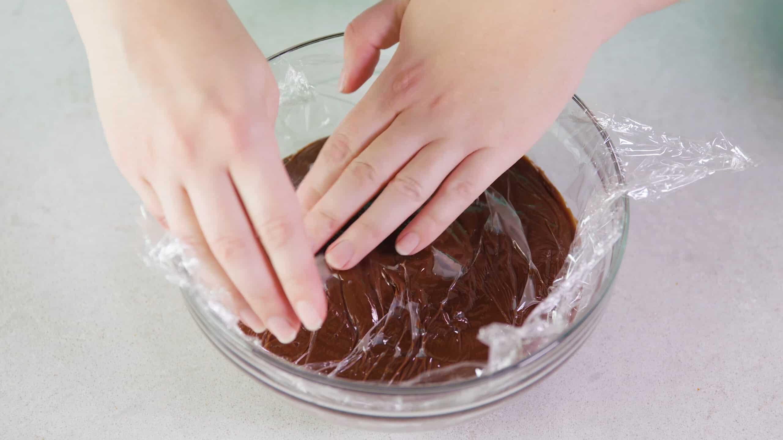 Overhead view of clear glass mixing bowl filled with chocolate pudding and clear plastic wrap smoothed over the top of the pudding to keep from exposure to air while in the fridge.