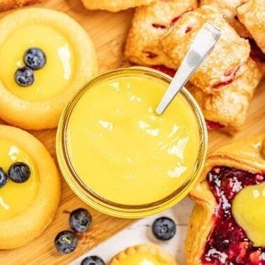 Bird's eye view of Lemon curd in a glass bowl with a spoon in it surrounded by various pastries.