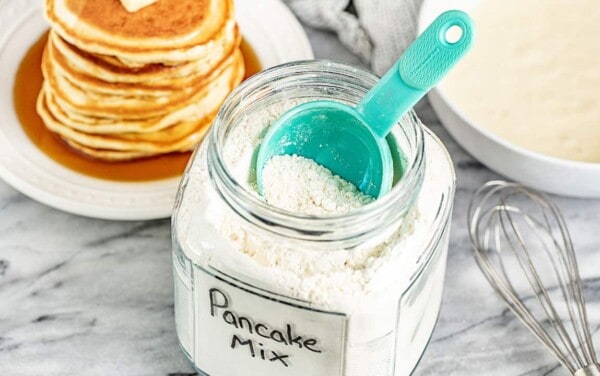 A glass container full of Pancake Mix with a measuring cup in it.