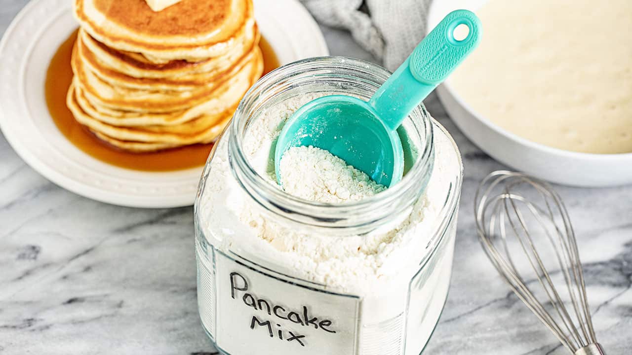 A glass container full of Pancake Mix with a measuring cup in it.