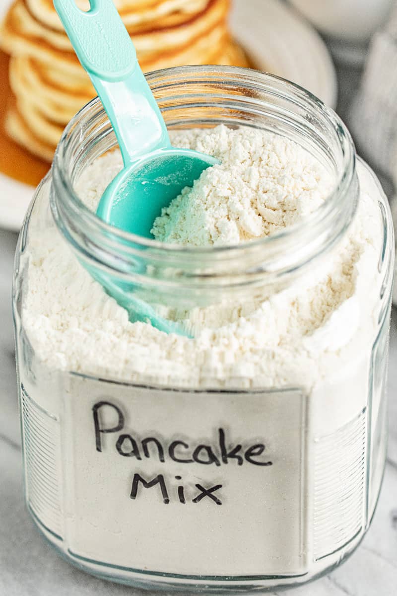 A glass container full of Pancake Mix with a measuring cup in it.