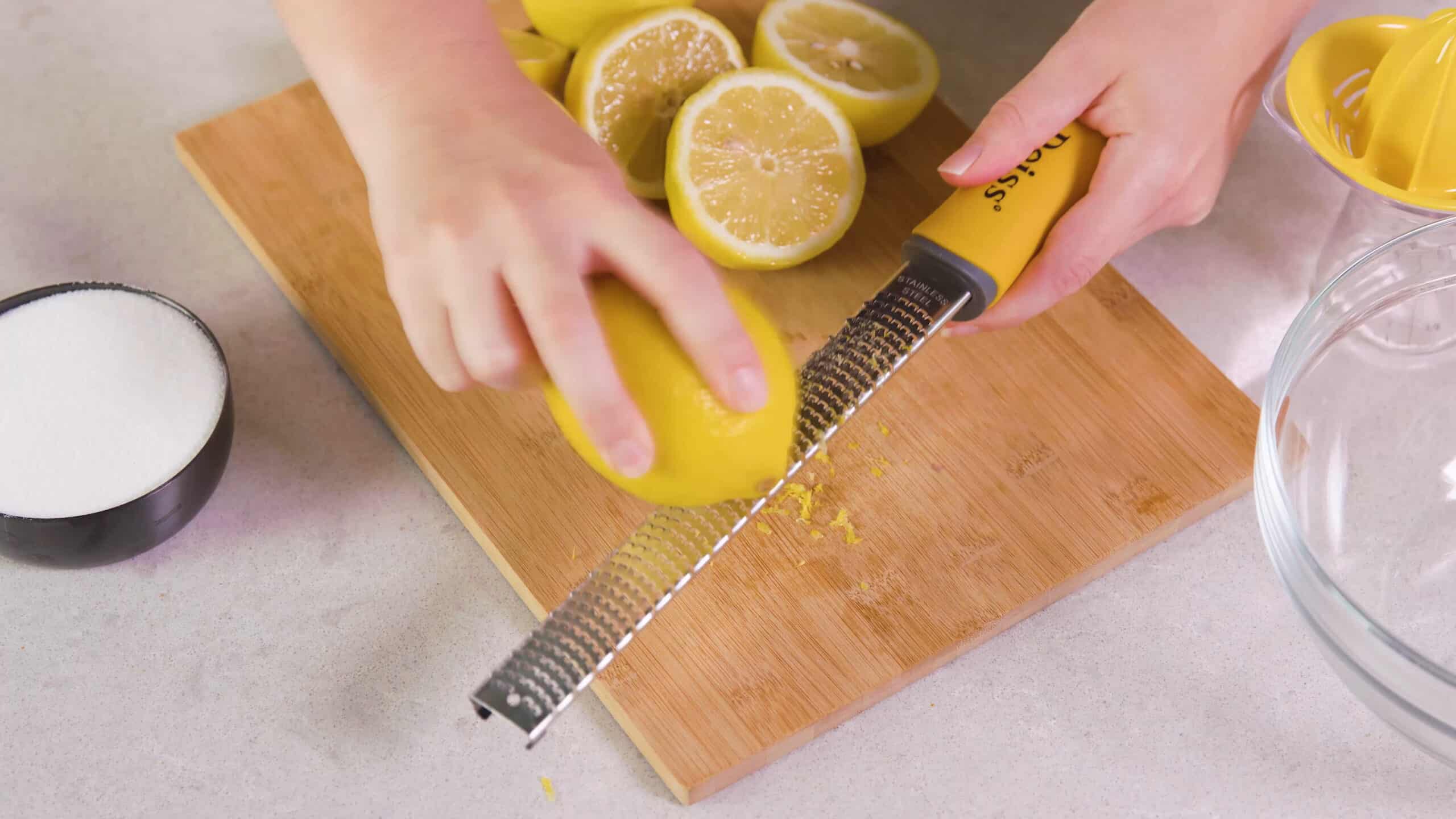 Overhead view of one lemon being zested with a stainless steel fruit zester on a cutting board with three halved lemons ready to juice.