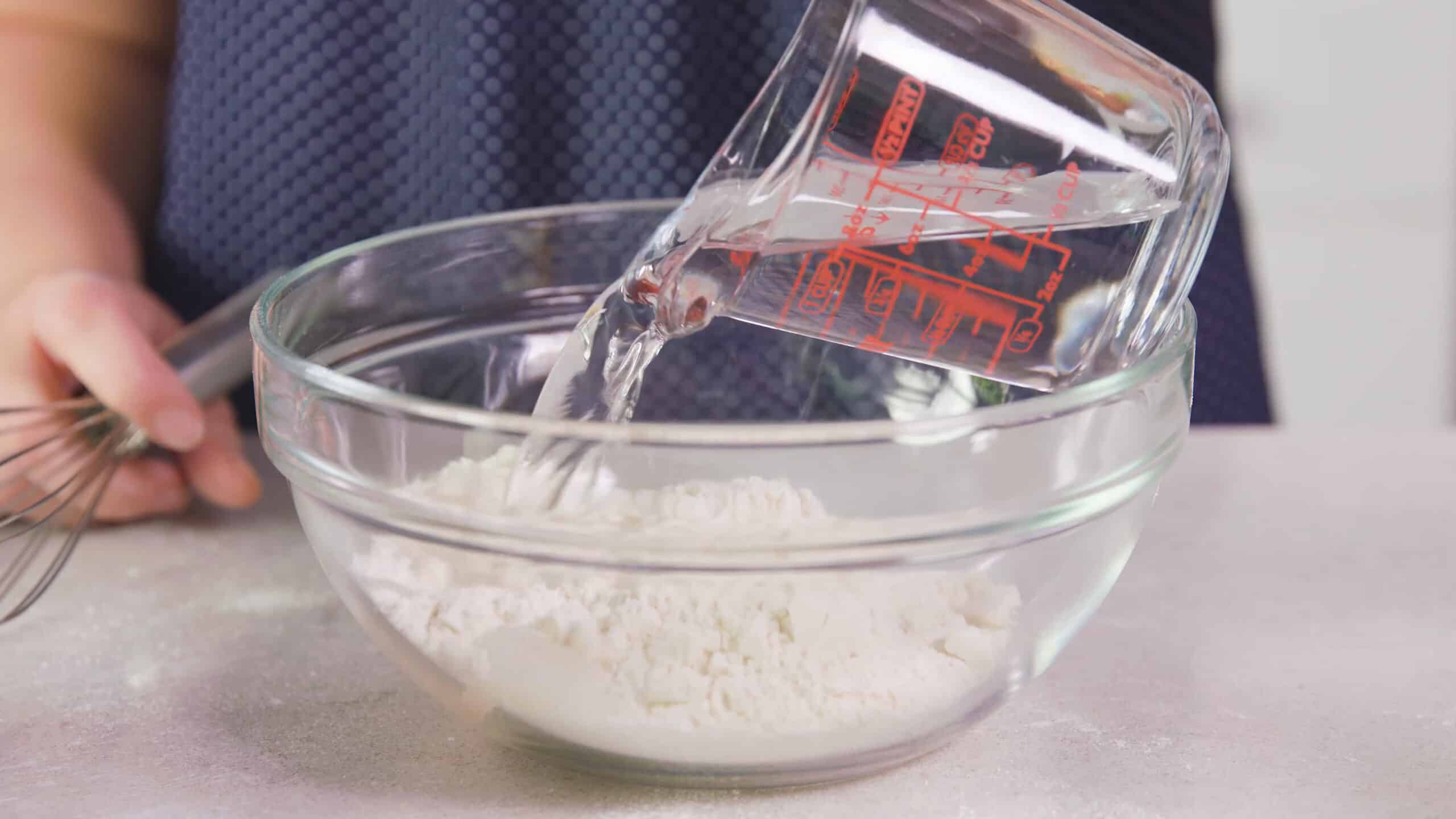 Side view of large clear mixing bowl with dry pancake mix and water being poured in from a clear measuring cup.