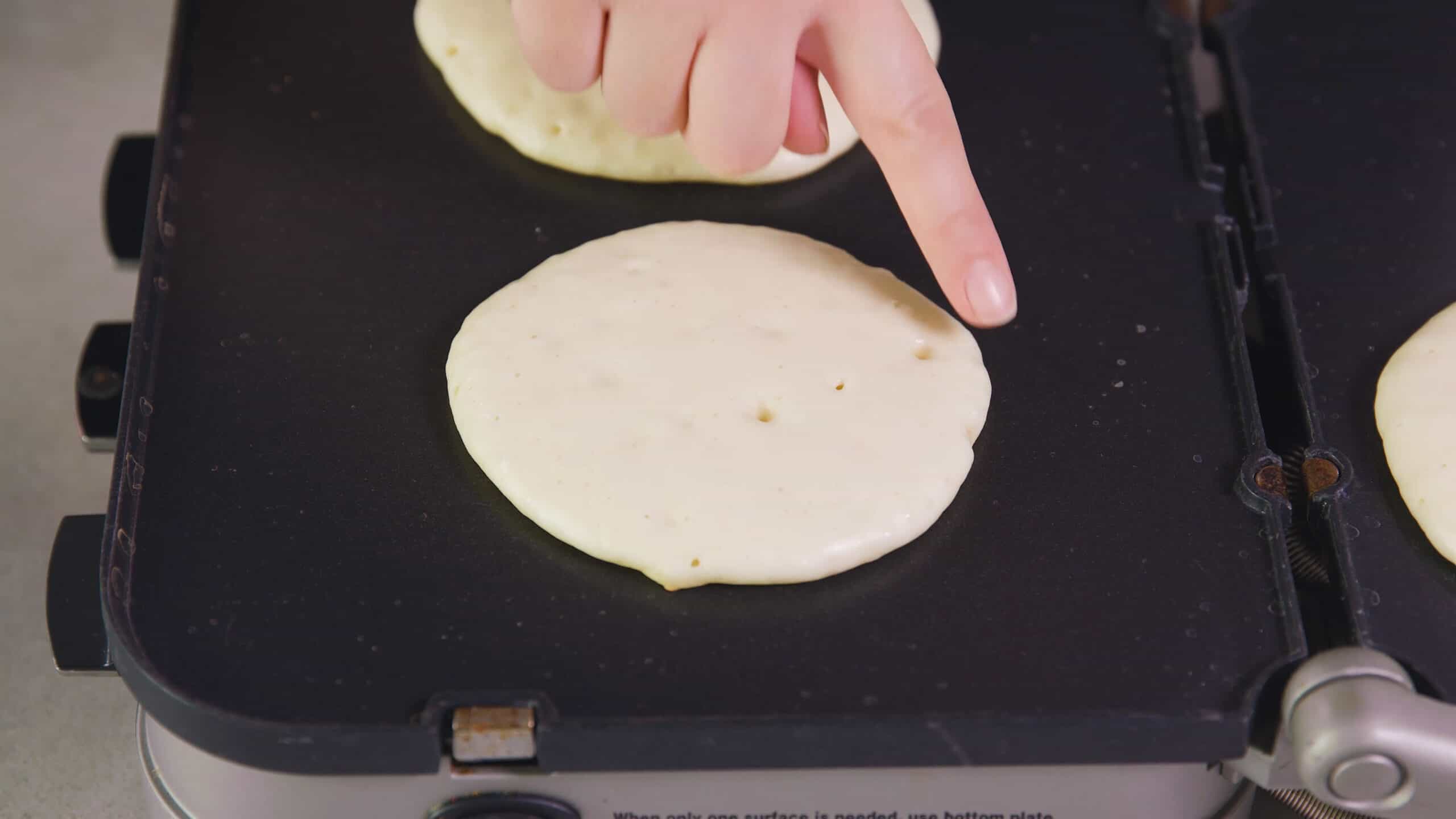 Angled view of non-stick griddle with a cooking pancake on top with the doughy side up and bubbles just beginning to form to show correct doneness.