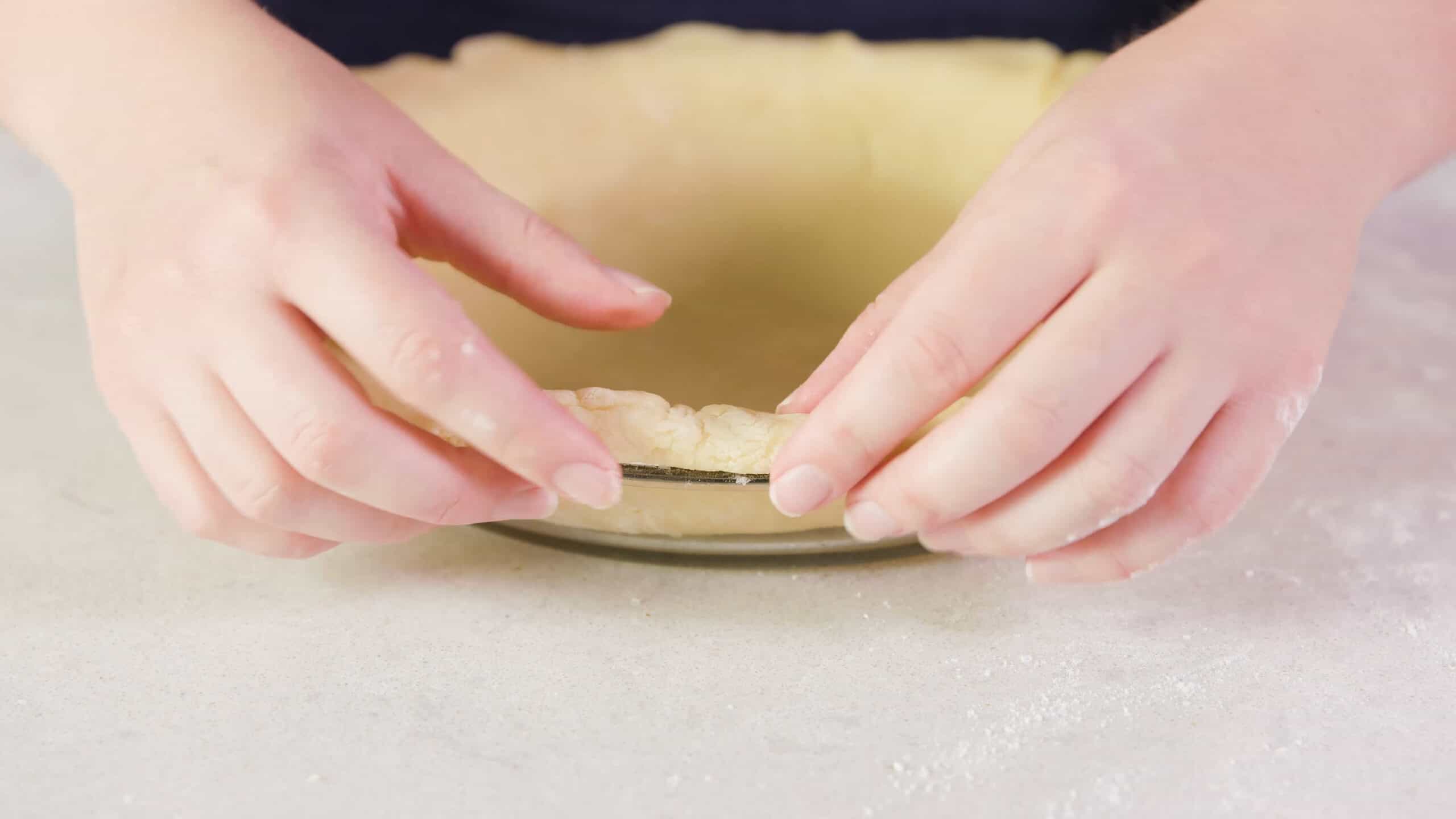 Close-up view of outer edge of clear glass pie plate with the rolled pie crust dough placed with a puckered edge ready for pleating.