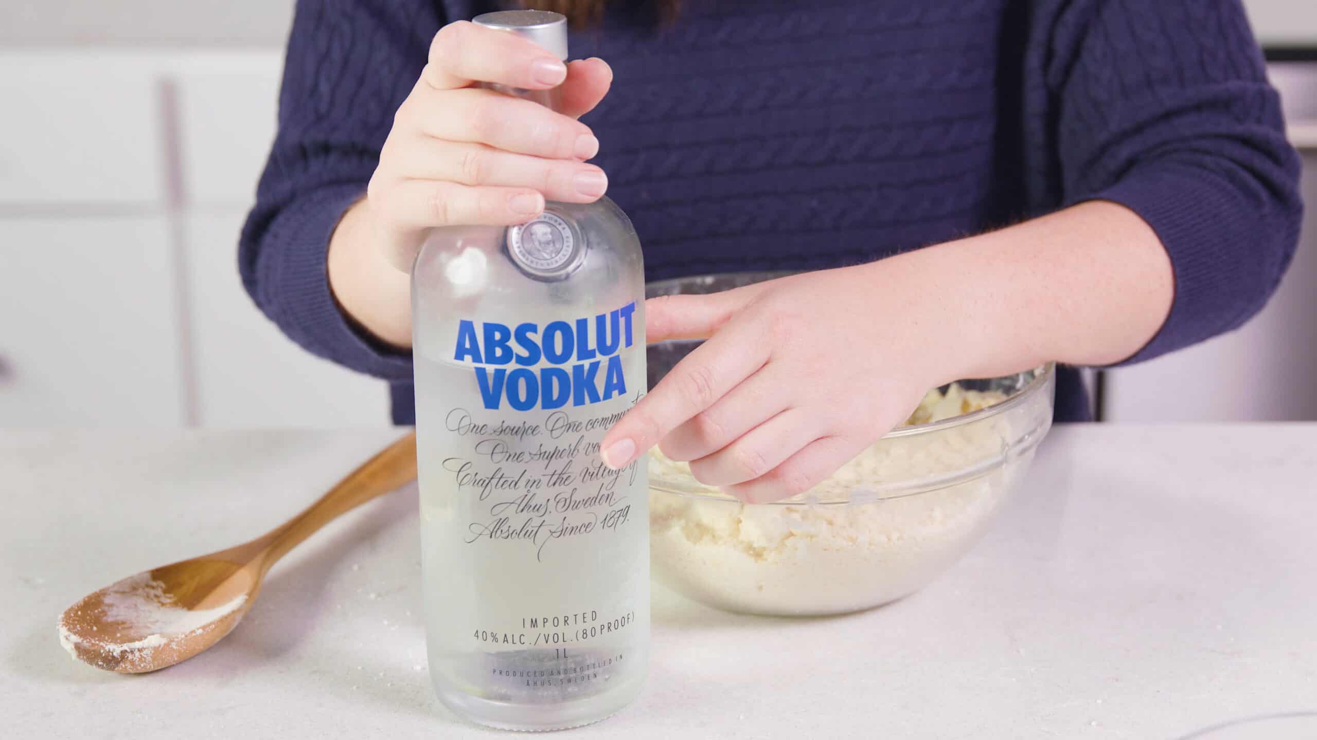 Angled view of a bottle of vodka with a large clear glass mixing bowl behind filled with pie crust dough all on a marble countertop.