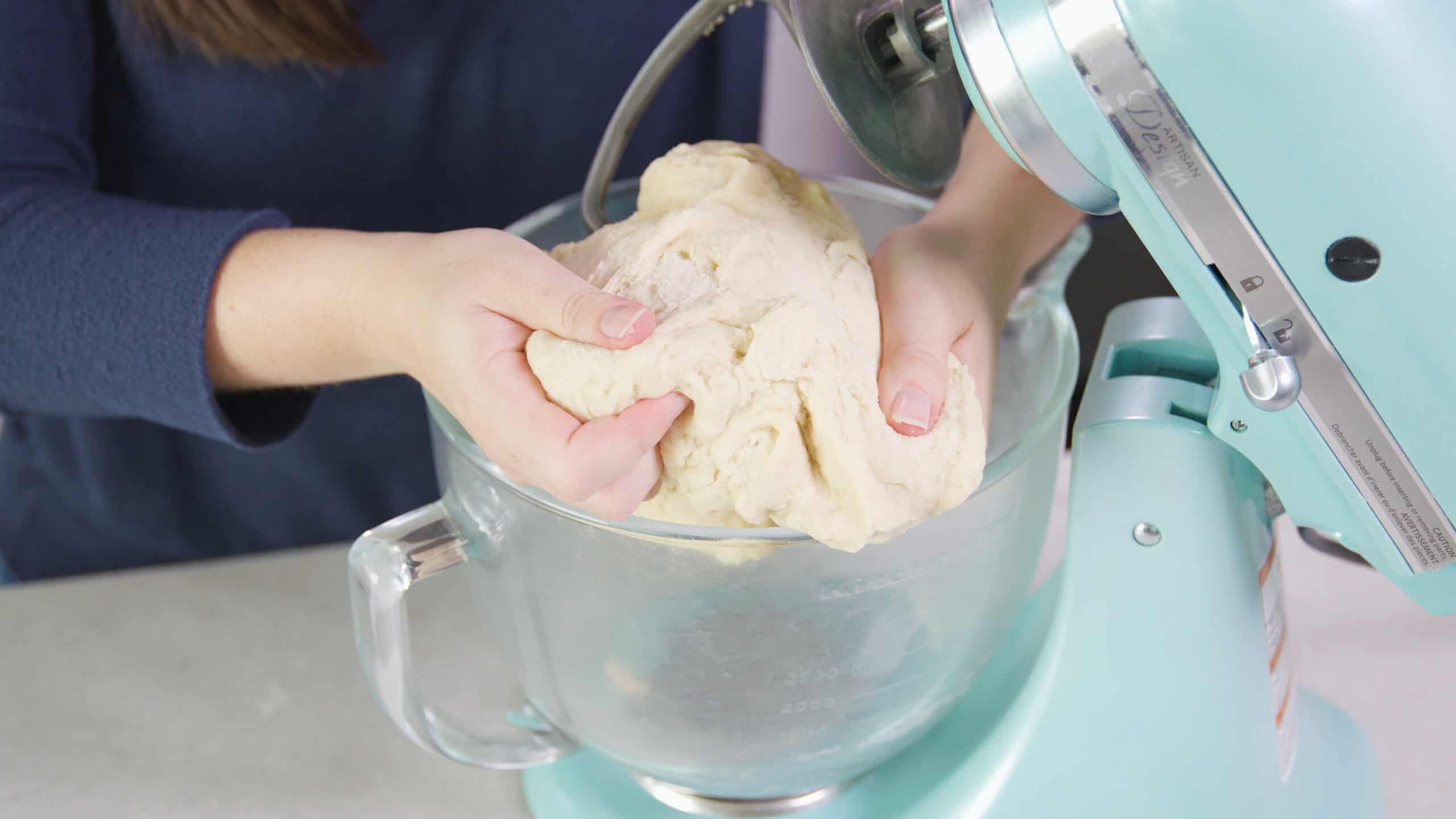 Angled view of clear glass mixing bowl attached to countertop mixer and the dough lifted to show texture of dough ready for rolling.