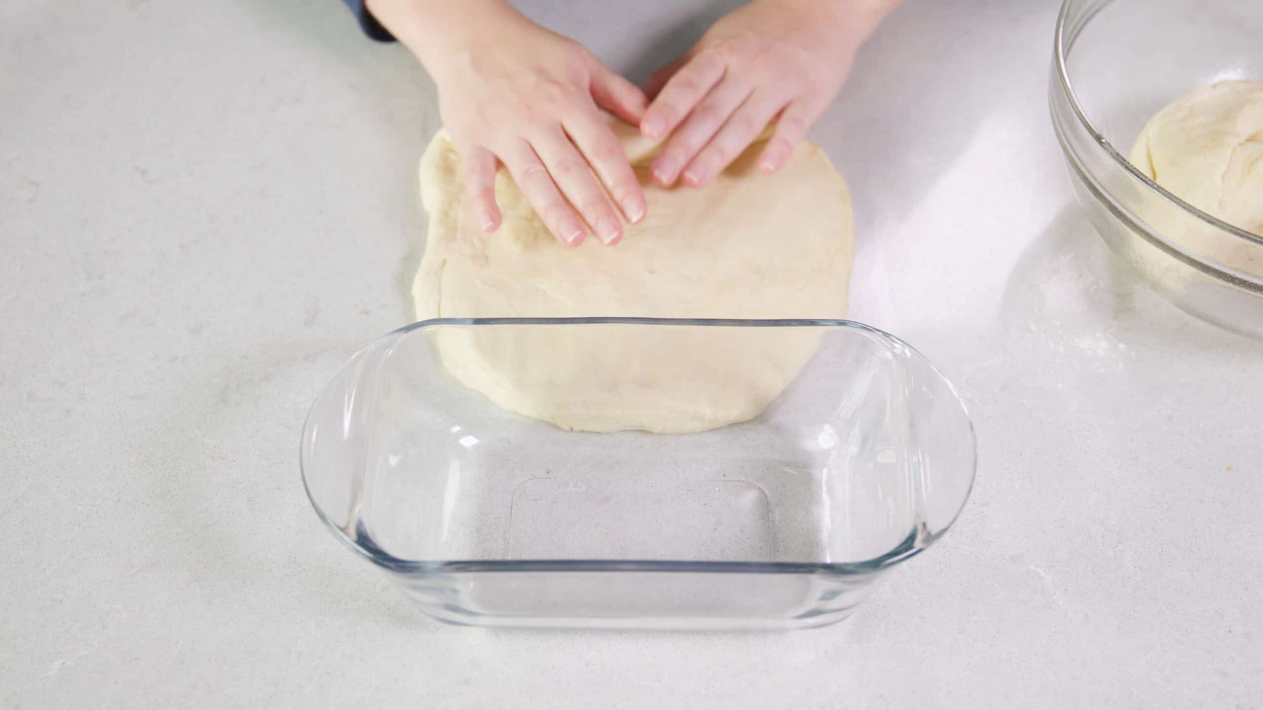 Overhead view of clean marble countertop with a clear glass bread pan used to reference correct width for the loaf in front of rolled out bread dough.
