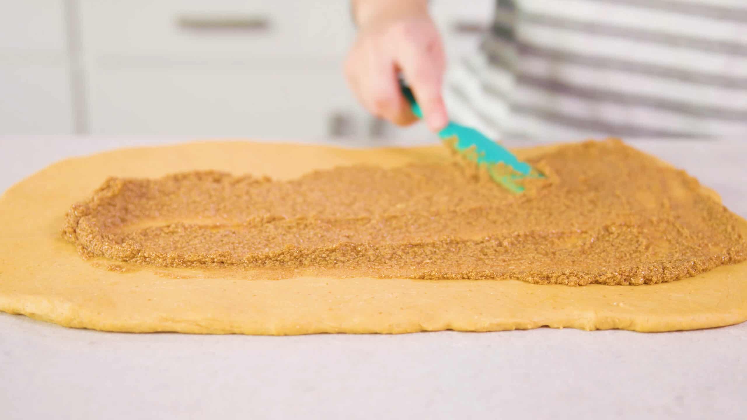 Angled view of pumpkin cinnamon roll mix spread onto the rolled out dough with a plastic spatula all on a clean marble countertop.