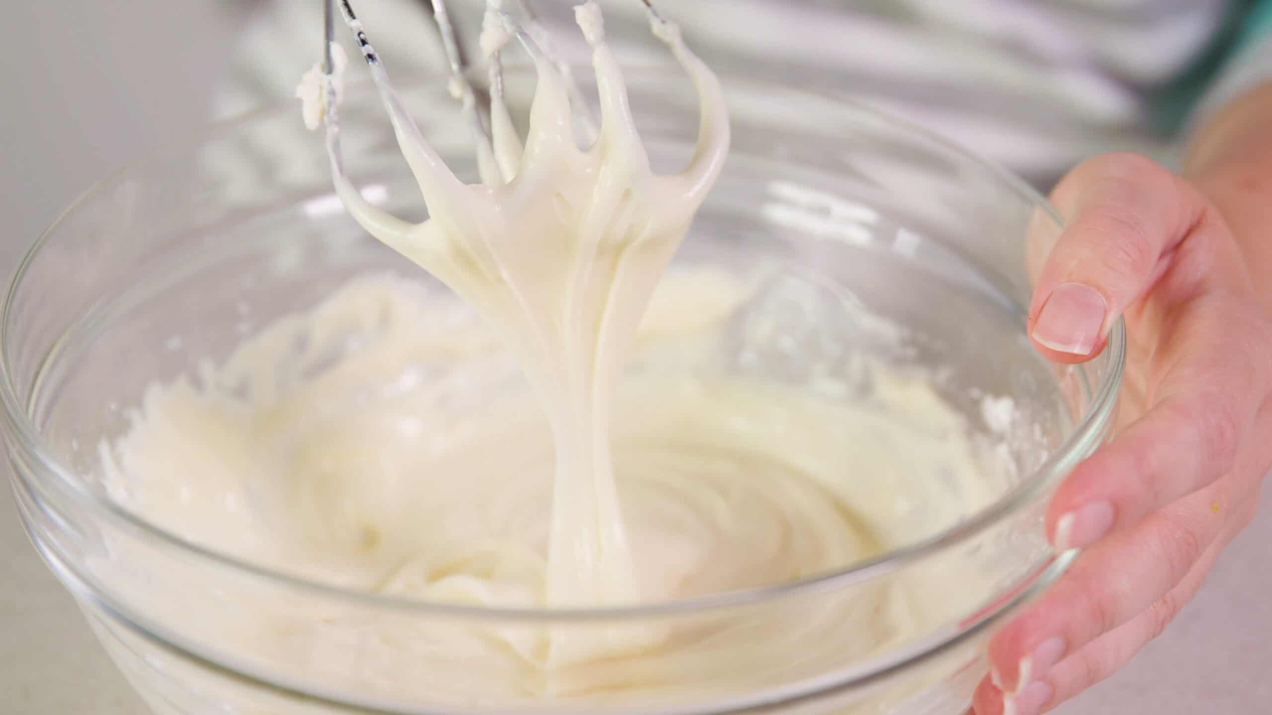 Close-up view of white cream cheese frosting on hand mixer whisks lifted from a large clear mixing bowl setting on a countertop.