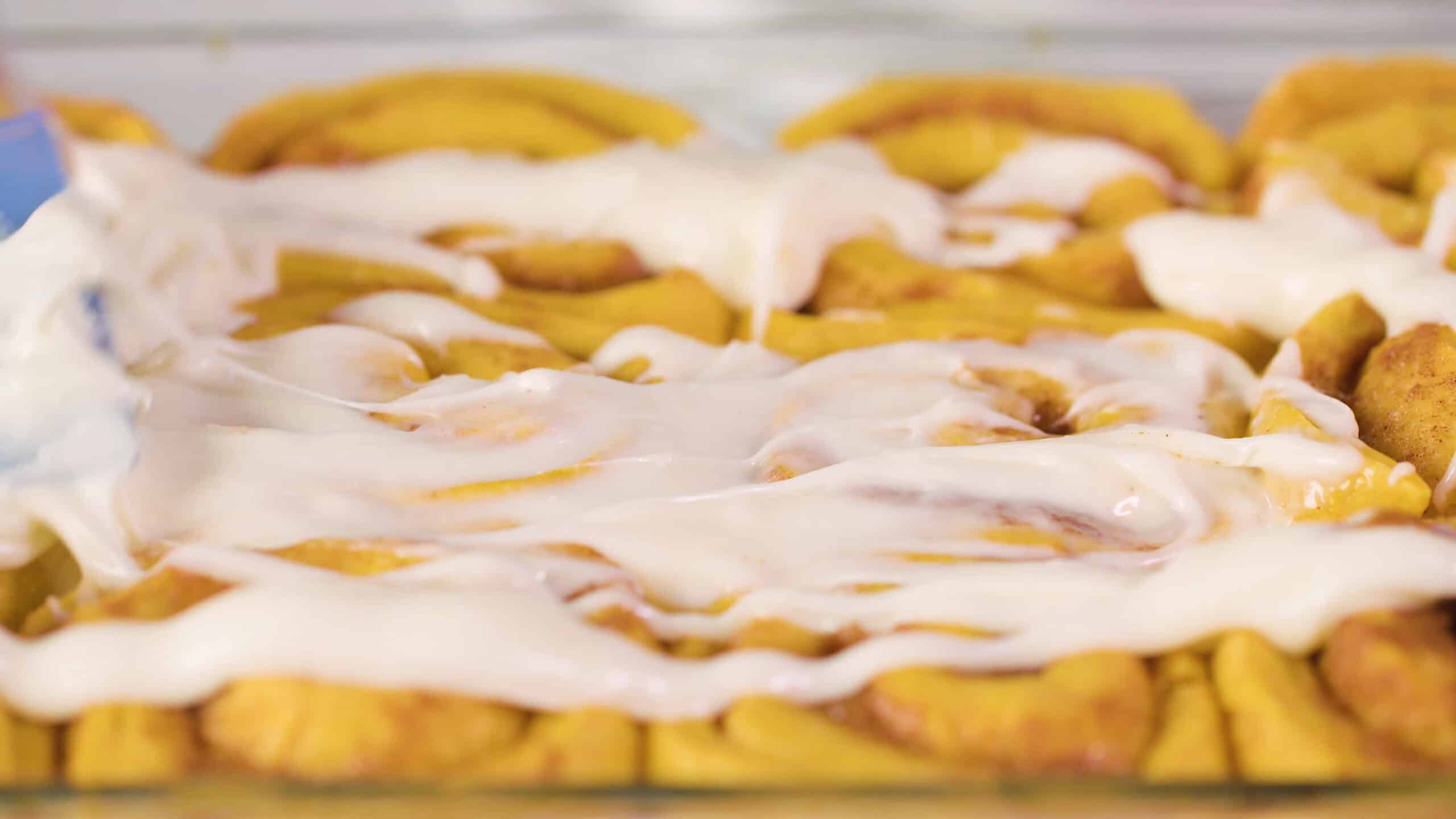 Close-up view of baked pumpkin cinnamon rolls still warm and white cream cheese frosting drizzled and spread on top with a plastic spatula.