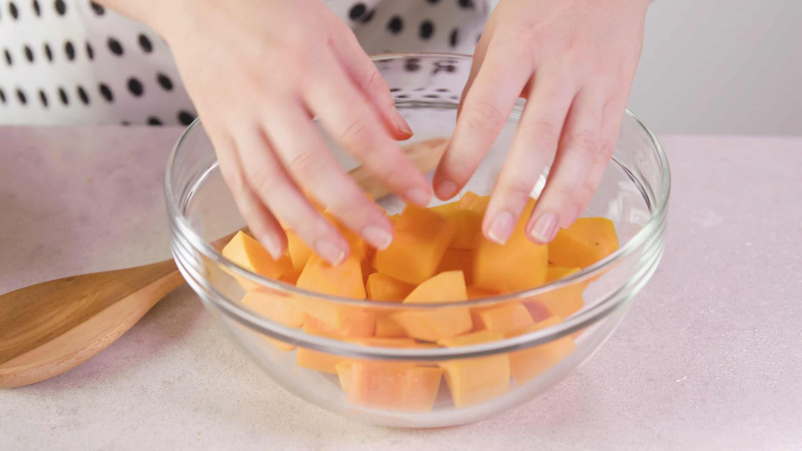 Angled view of large sized clear mixing bowl filled with diced butternut squash with a wooden spoon for mixing all on a marble countertop.