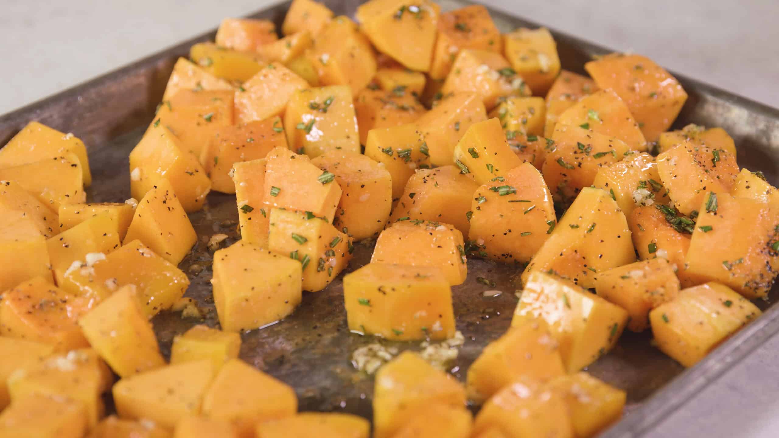 Close-up view of rosemary, garlic, and olive oil flavored butternut squash laying on a silver baking sheet.