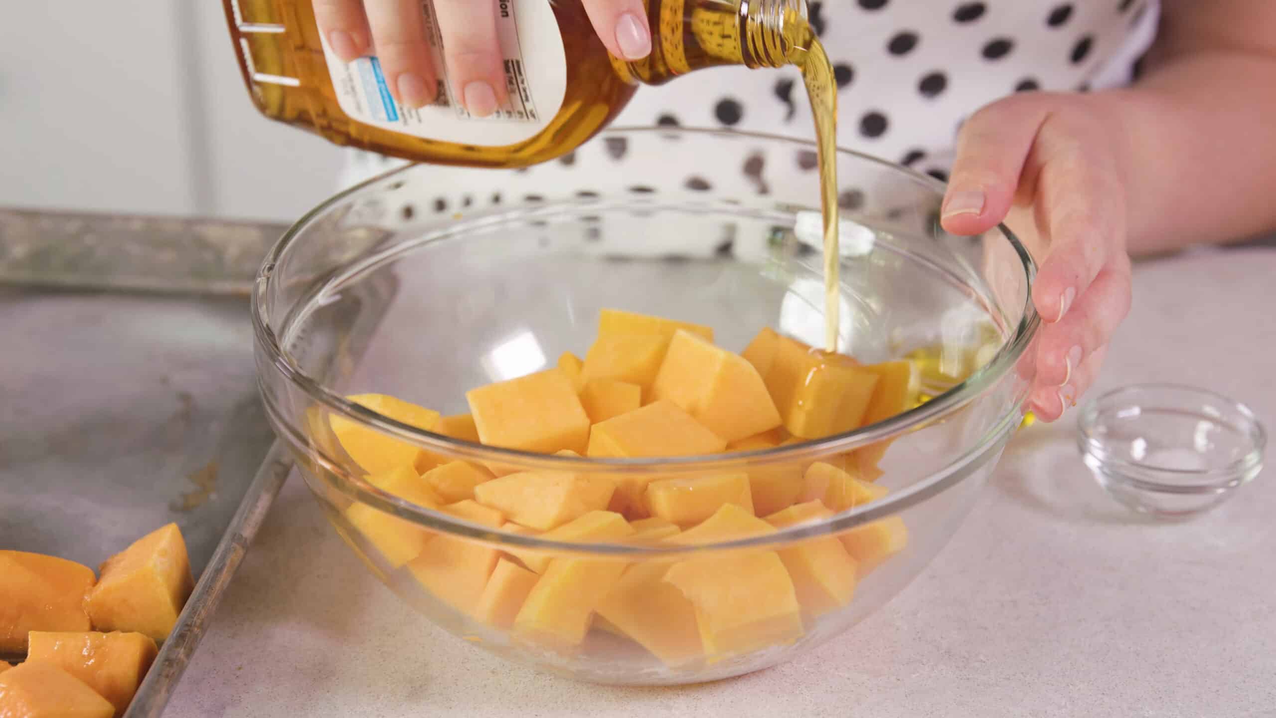 Angled view of clear mixing bowl filled with diced butternut squash and a bottle of maple syrup pouring over the top of the squash for flavoring.