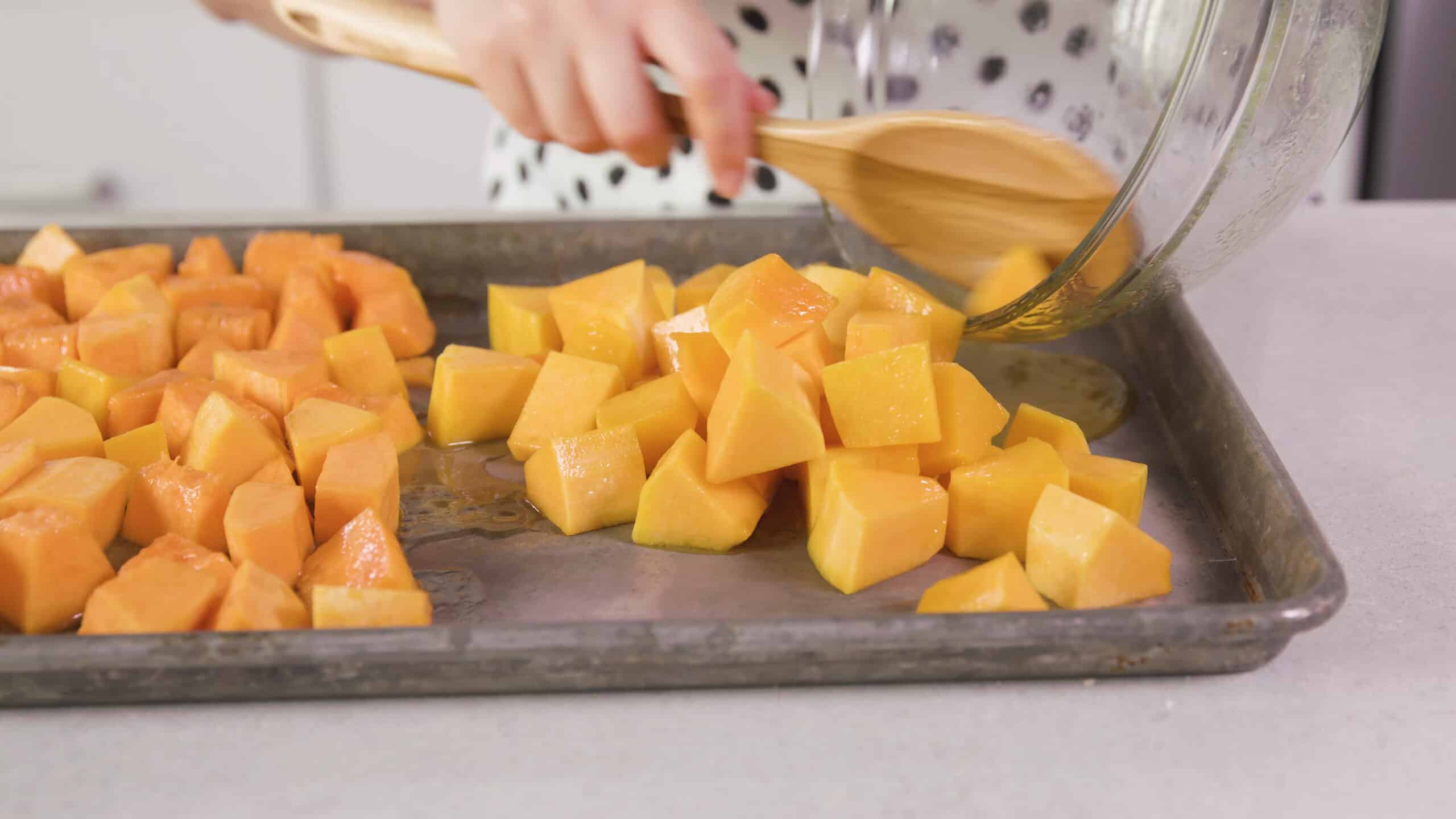 Angled view of silver baking sheet with maple flavored butternut squash being poured out from a clear mixing bowl using a wooden spoon.