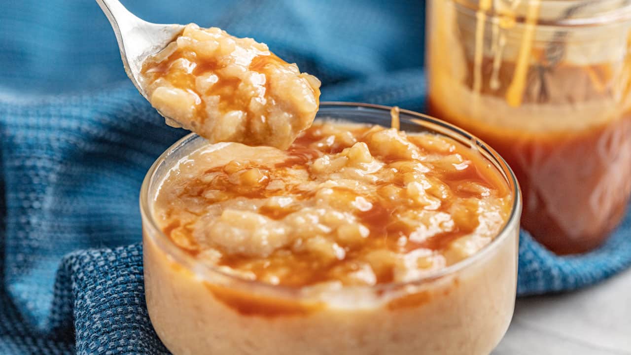 A spoon full of Salted Caramel Rice Pudding above a glass dish full of rice pudding drizzled with caramel sauce.