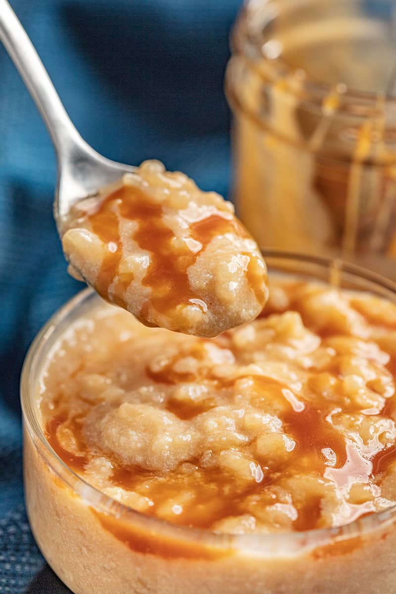 A spoon full of Salted Caramel Rice Pudding above a glass dish full of rice pudding.