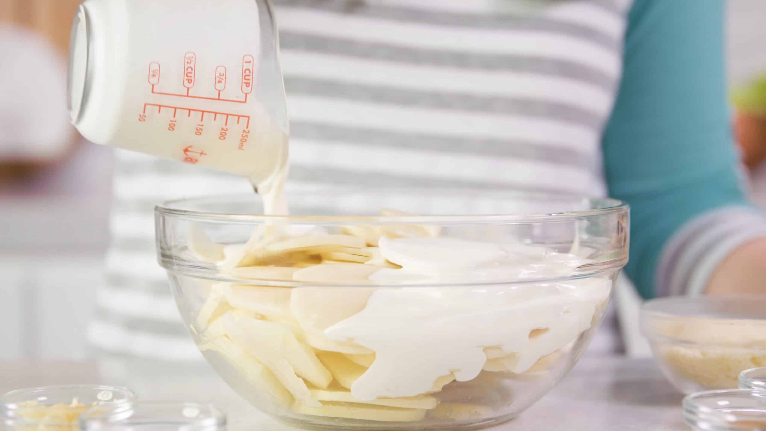 Side view of large glass mixing bowl filled with thinly sliced russet potatoes and heavy cream being poured on top from a clear glass measuring cup.