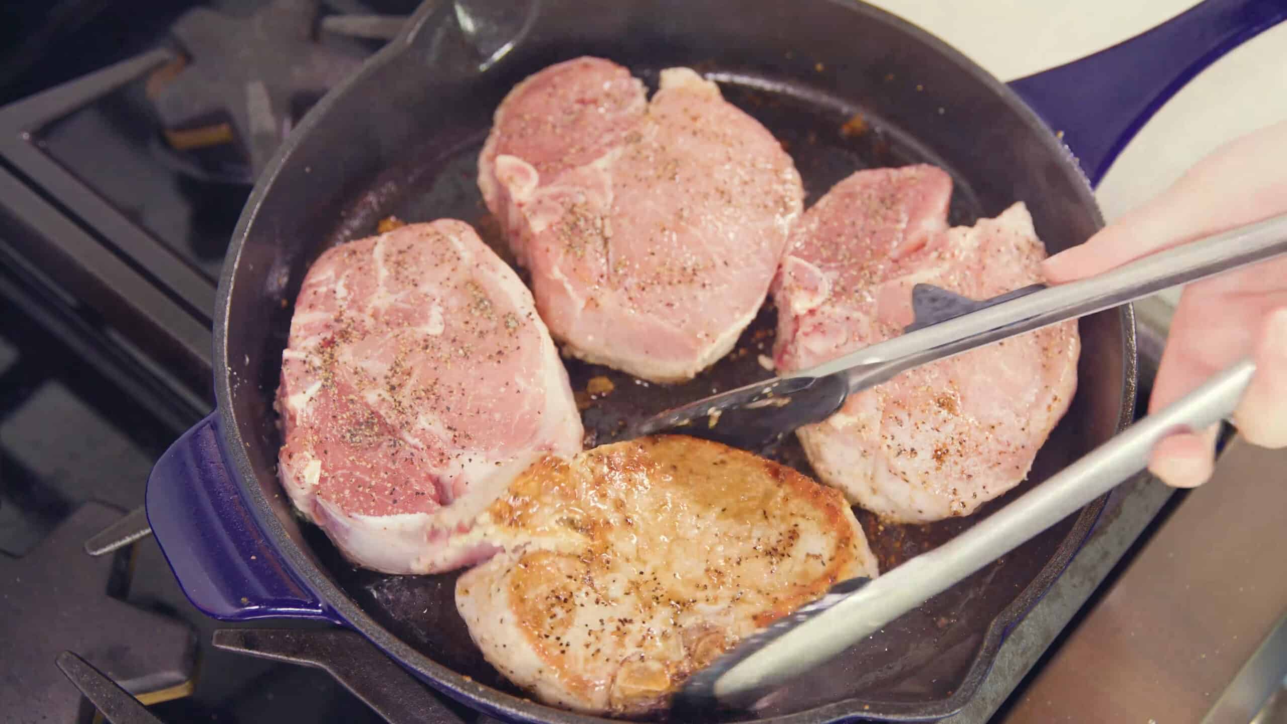 Overhead view of blue enamel cast iron skillet with four pork chops, one which has been flipped to show the seared side up.