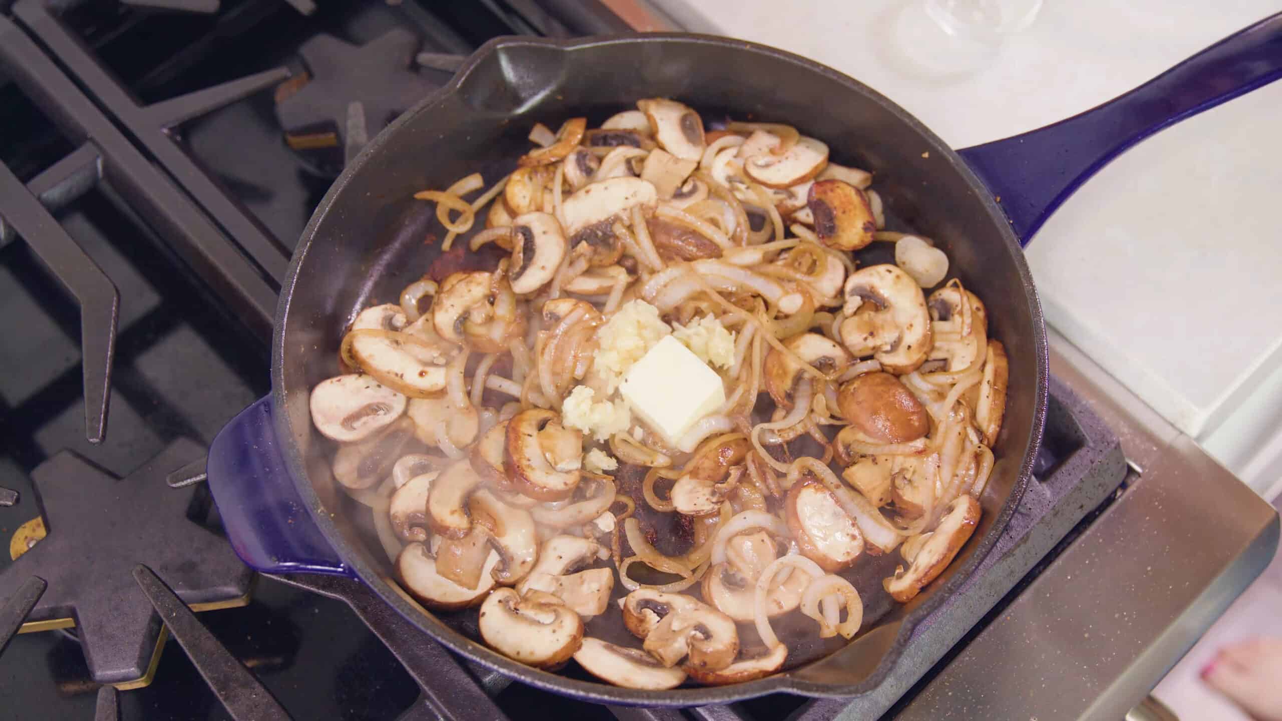 Overhead view of blue enamel cast iron skillet with sautéed mushrooms and onions with minced garlic and a pad of butter beginning to melt in the middle.
