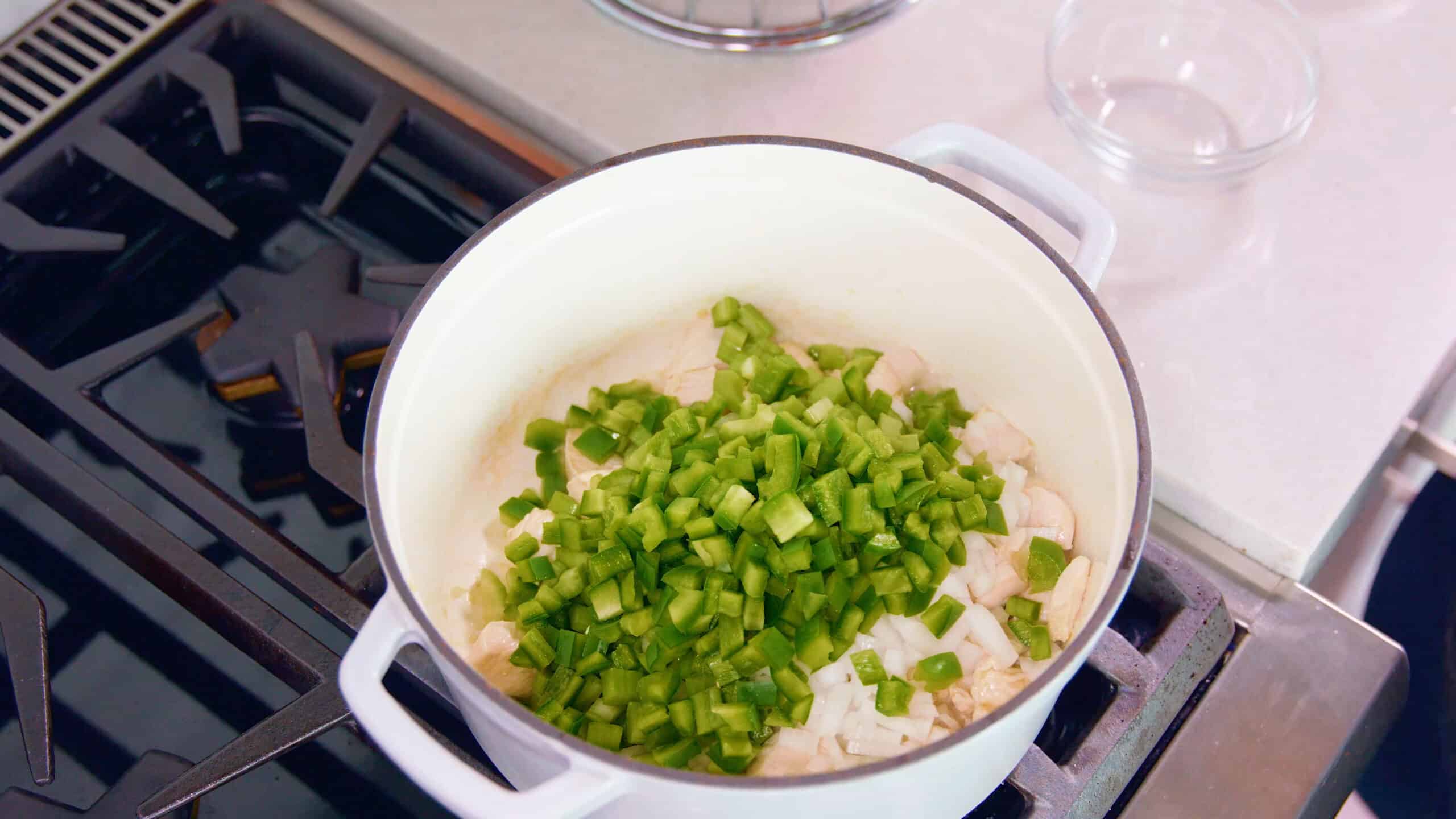 Overhead view of white enamel cast iron pot with green peppers and white onion added to cooked chicken breast on a stovetop.