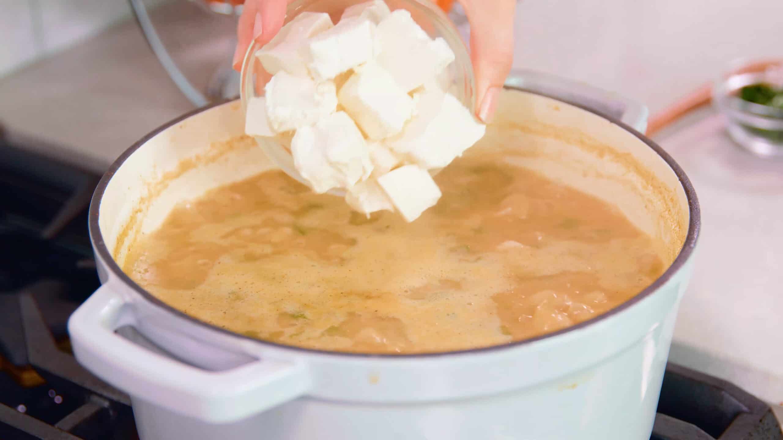 Angled view of white enamel cast iron pot on the stovetop with cubes of cream cheese being added from a clear glass bowl