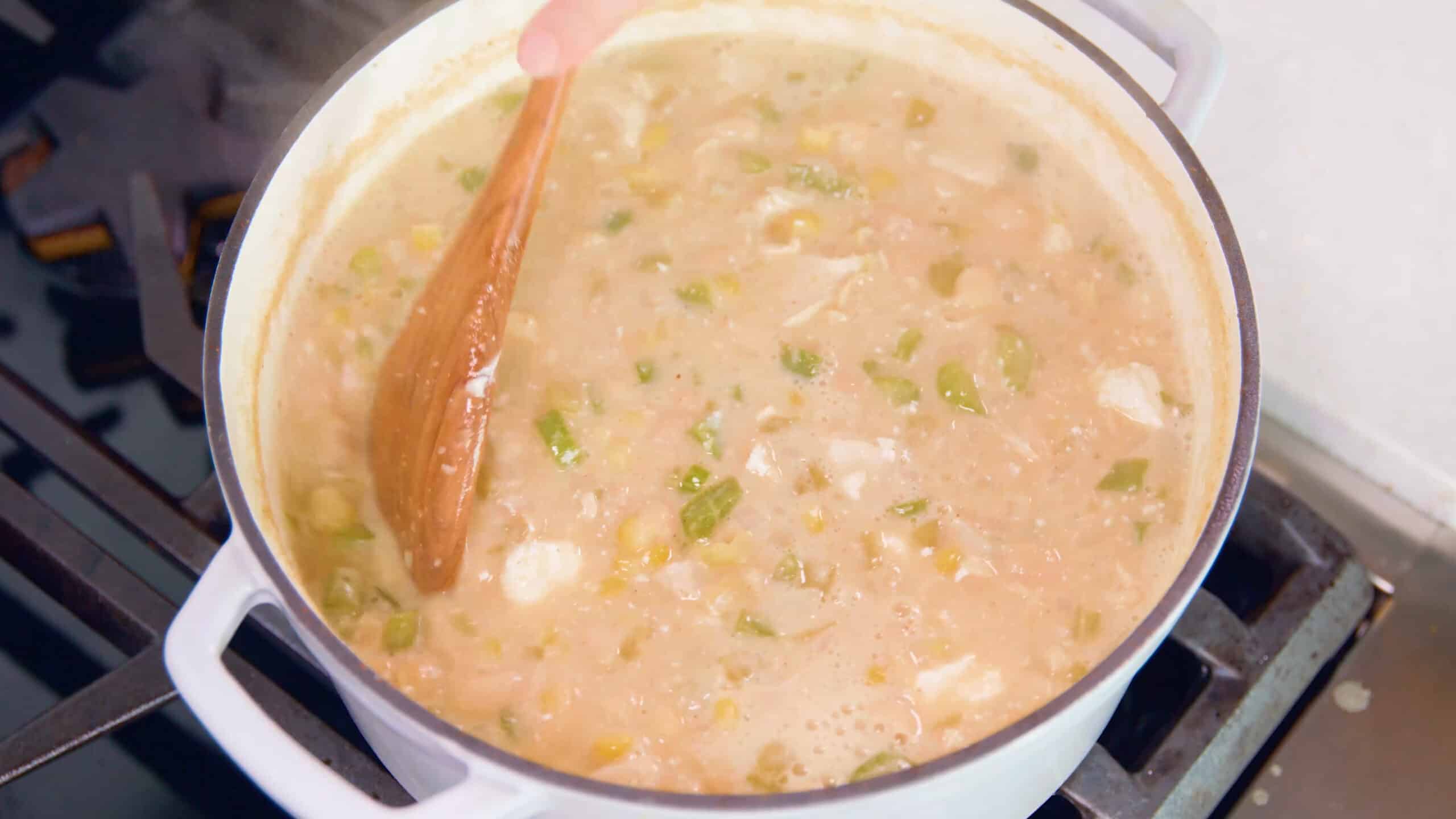 Overhead view of white enamel cast iron pot filled with white chicken chili simmers on the stovetop while a wooden spatula stirs the ingredients together.