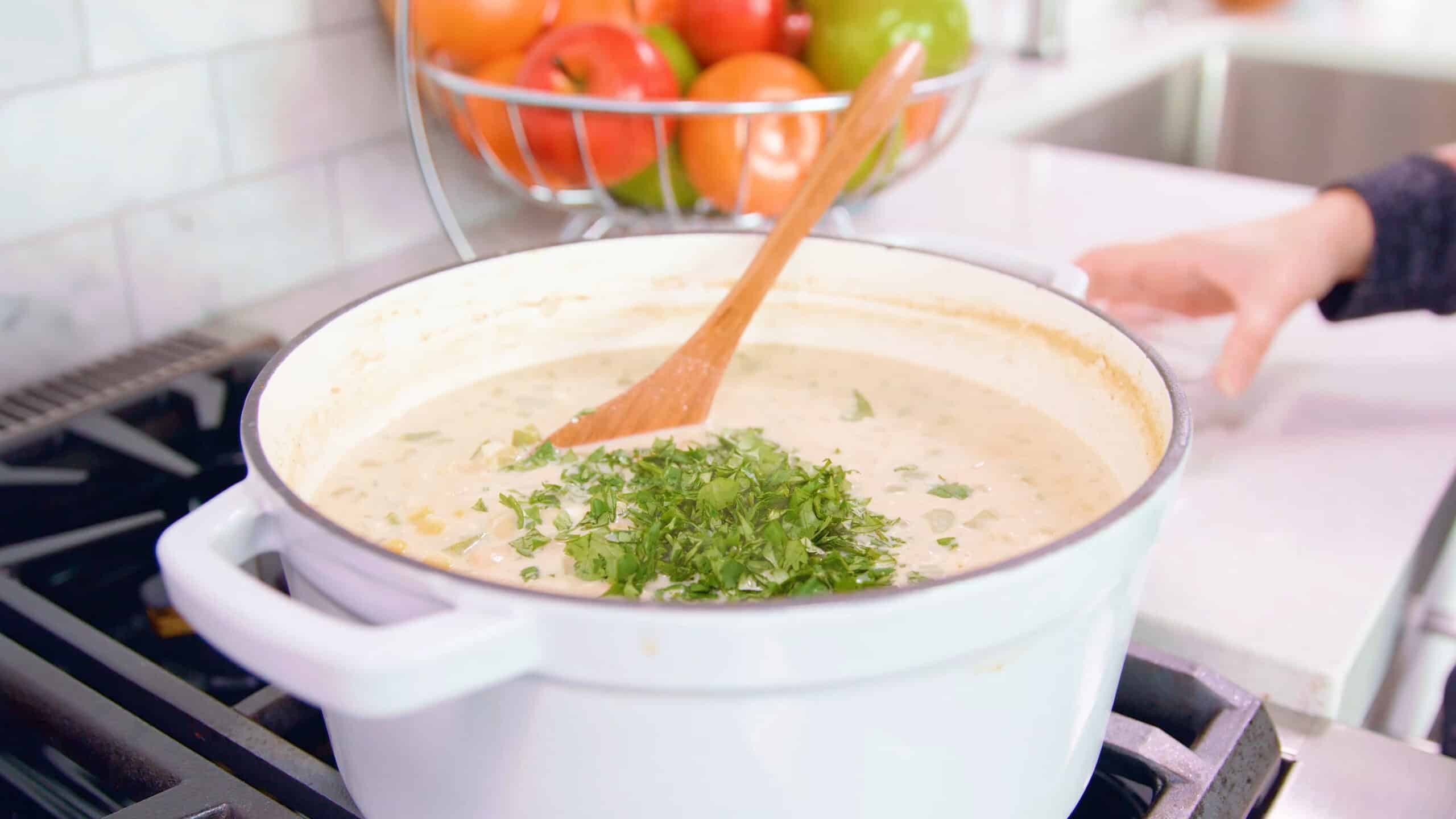 Angled view of white enamel cast iron pot filled with white chicken chili and finely chopped cilantro resting on top with a wooden spatula ready to stir together all on a stovetop.