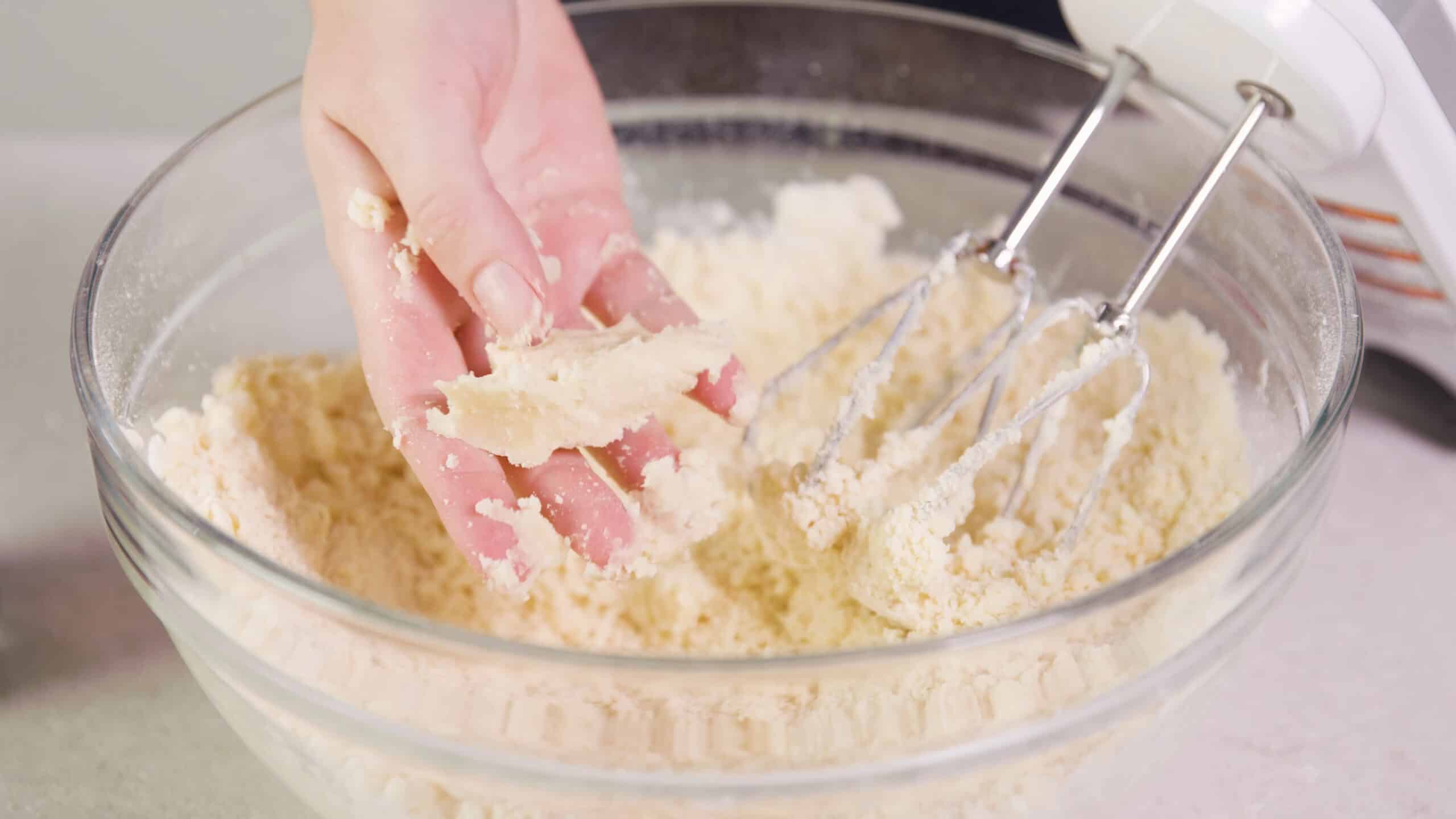 Close-up view of a large clear glass mixing bowl filled with shortbread cookies batter and a hand holding a portion of the batter that has been squeezed to show the texture once it has been pressed.