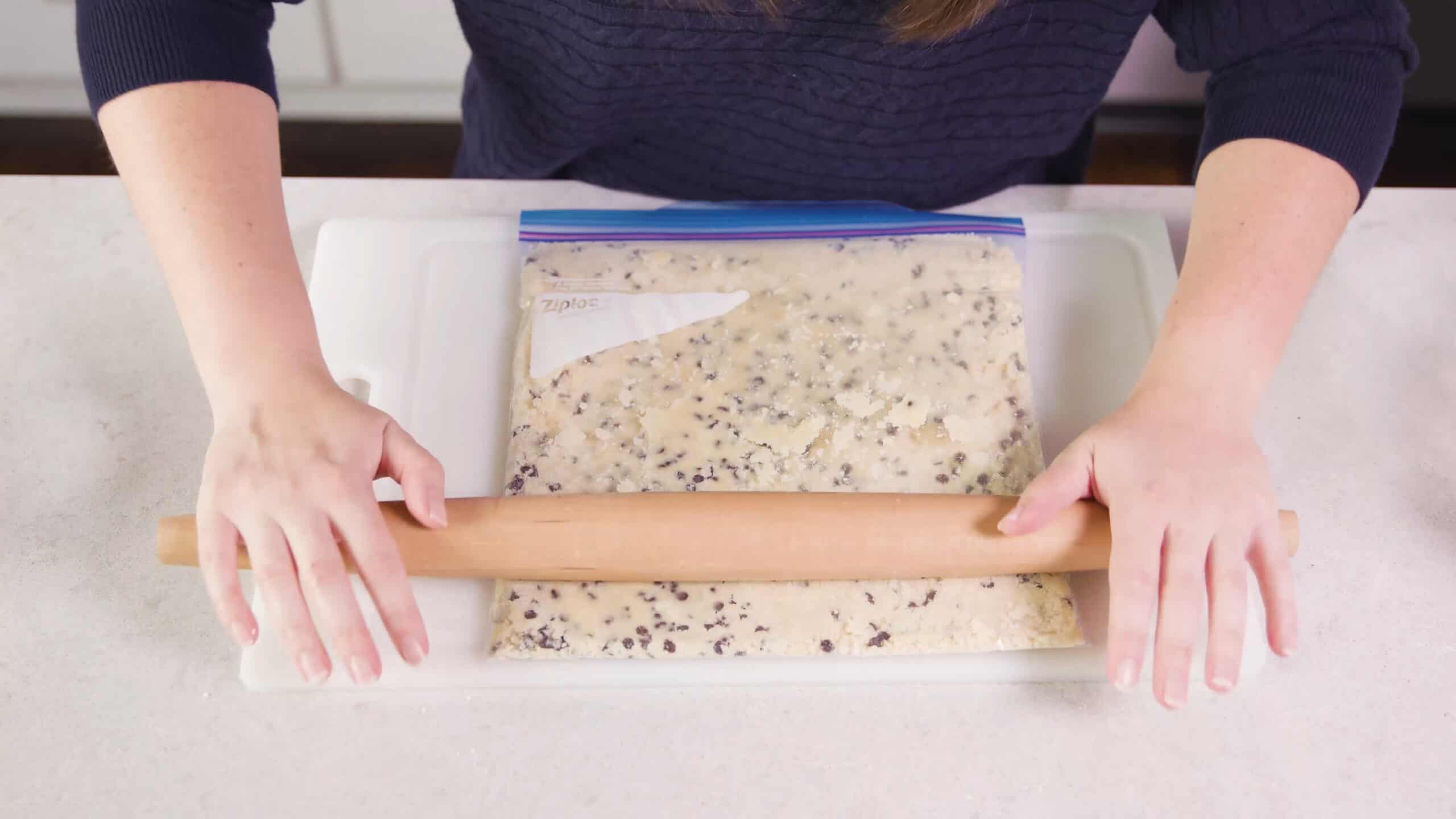 Overhead view of a clear plastic bag filled with chocolate chip shortbread cookie batter and being rolled with a wooden rolling pin on a white plastic cutting board all on a marble countertop.