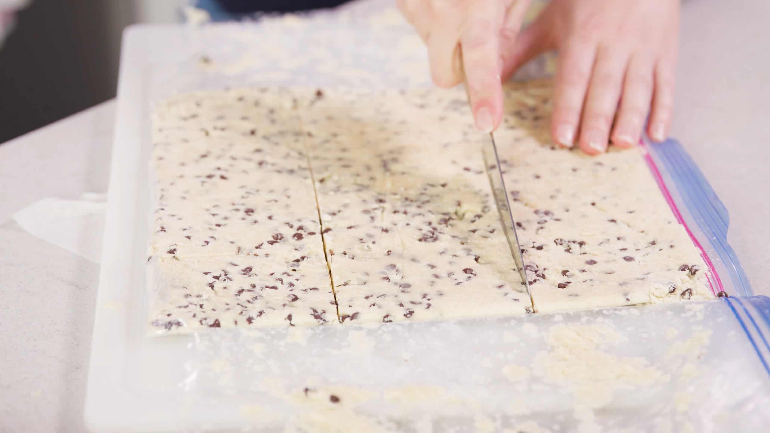 Angled view of a cut open clear plastic bag revealing the chocolate chip shortbread cookie dough and a kitchen knife cutting the dough into even sized rectangles.