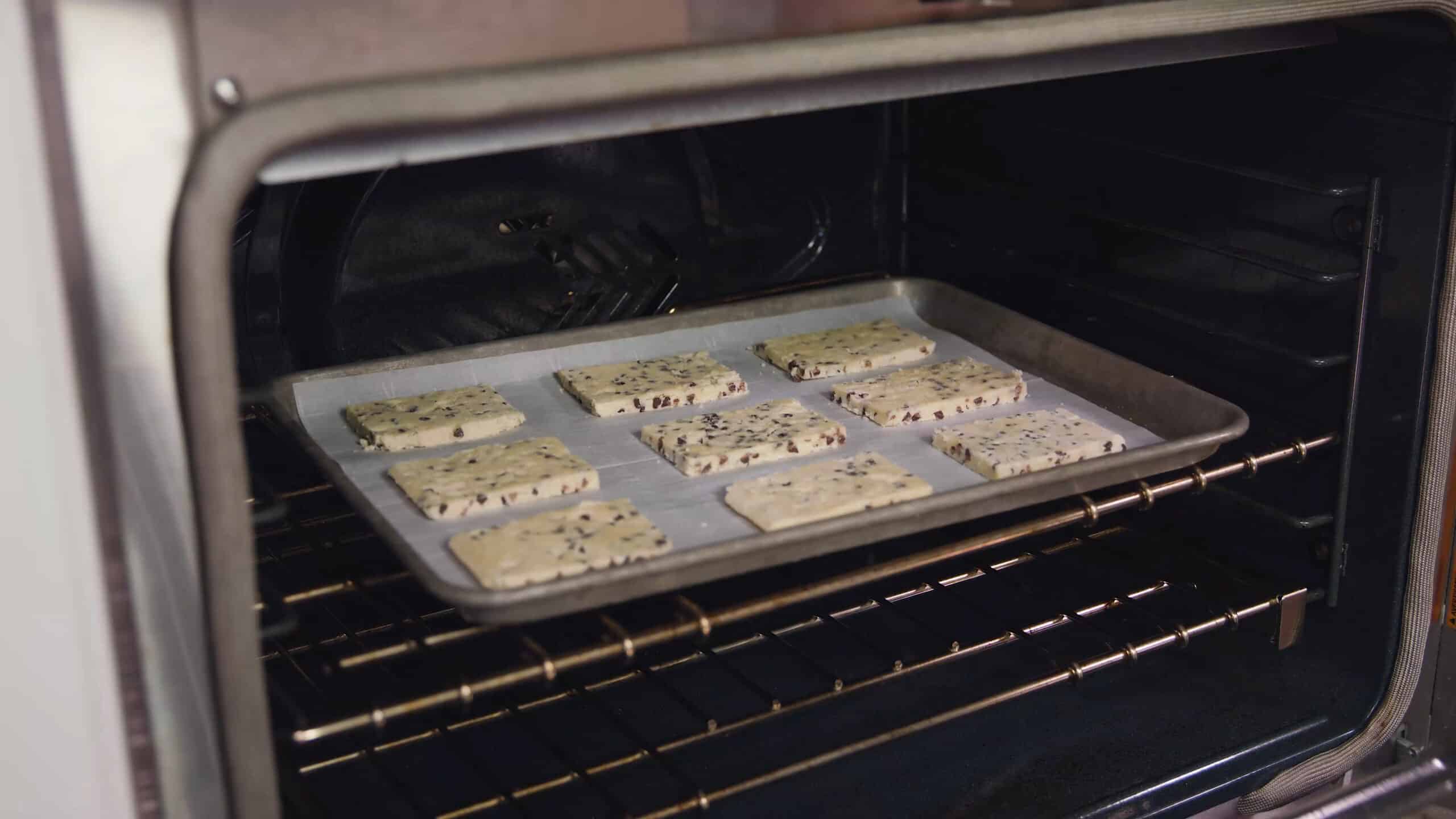 Angled view of an open oven with a metal baking sheet lined with parchment paper topped with nine chocolate chip shortbread cookies all on a metal rack in the middle of the oven.