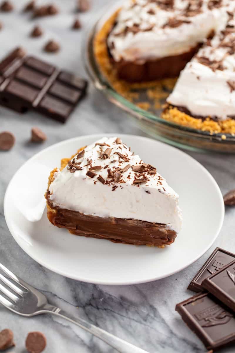 Slice of chocolate cream pie on a white plate with a chocolate pie in the background. 