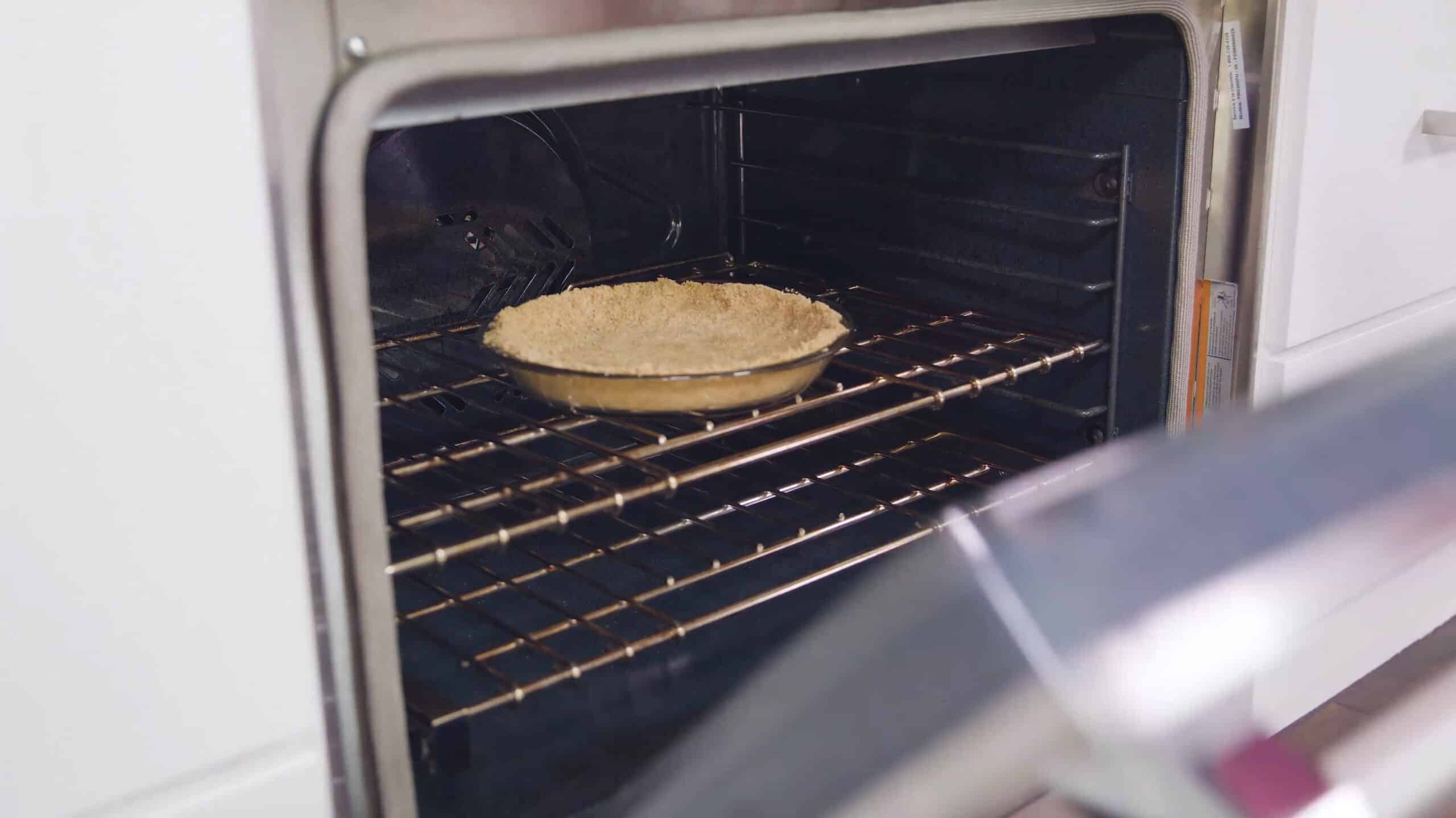 Angled view of open oven with clear glass pie plate with formed graham cracker crust inside placed on a rack in the middle of the oven.