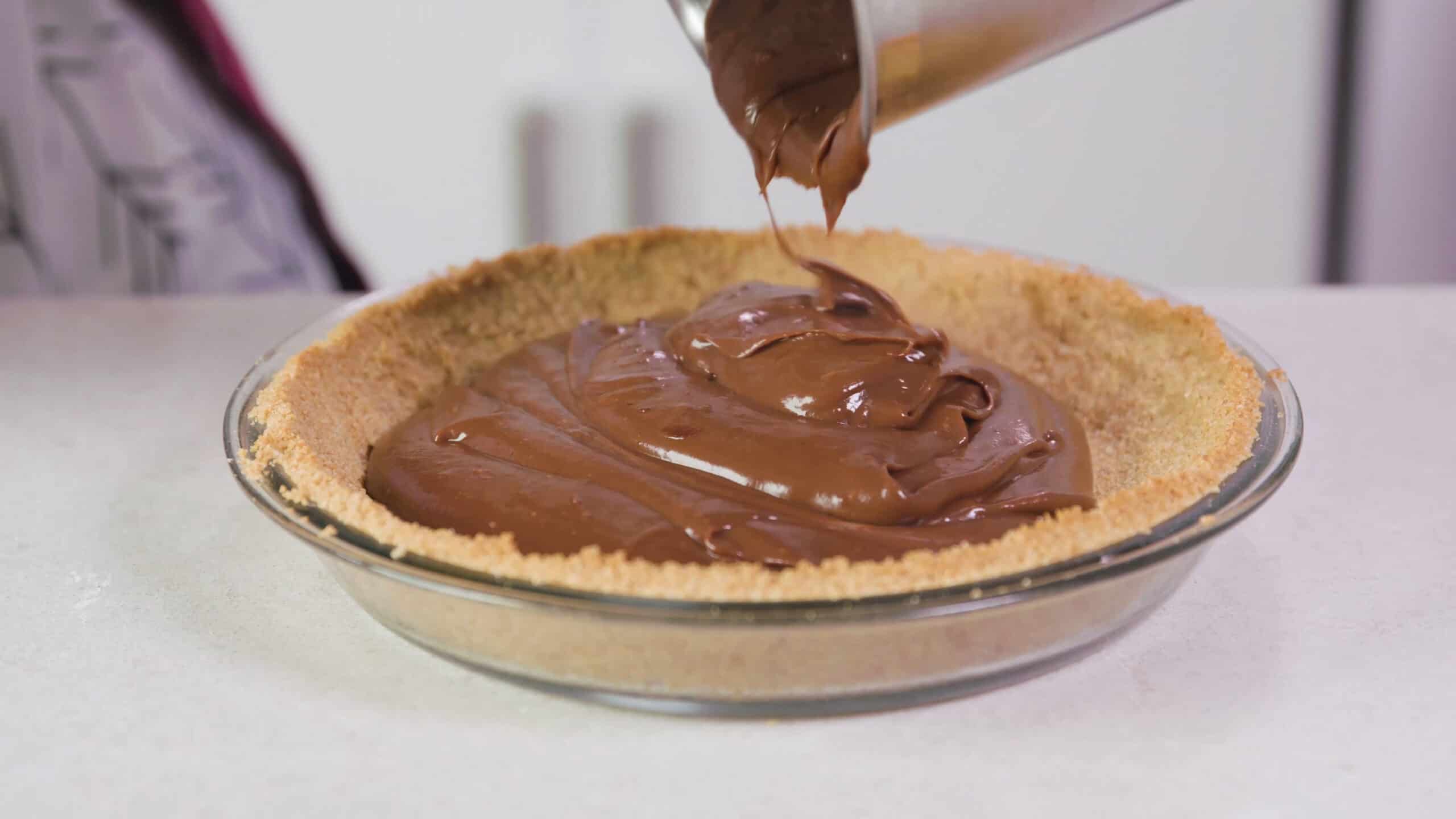 Angled view of cooked and cooled graham cracker pie crust in a clear glass pie plate and the chocolate filling being poured from a silver pot on top.