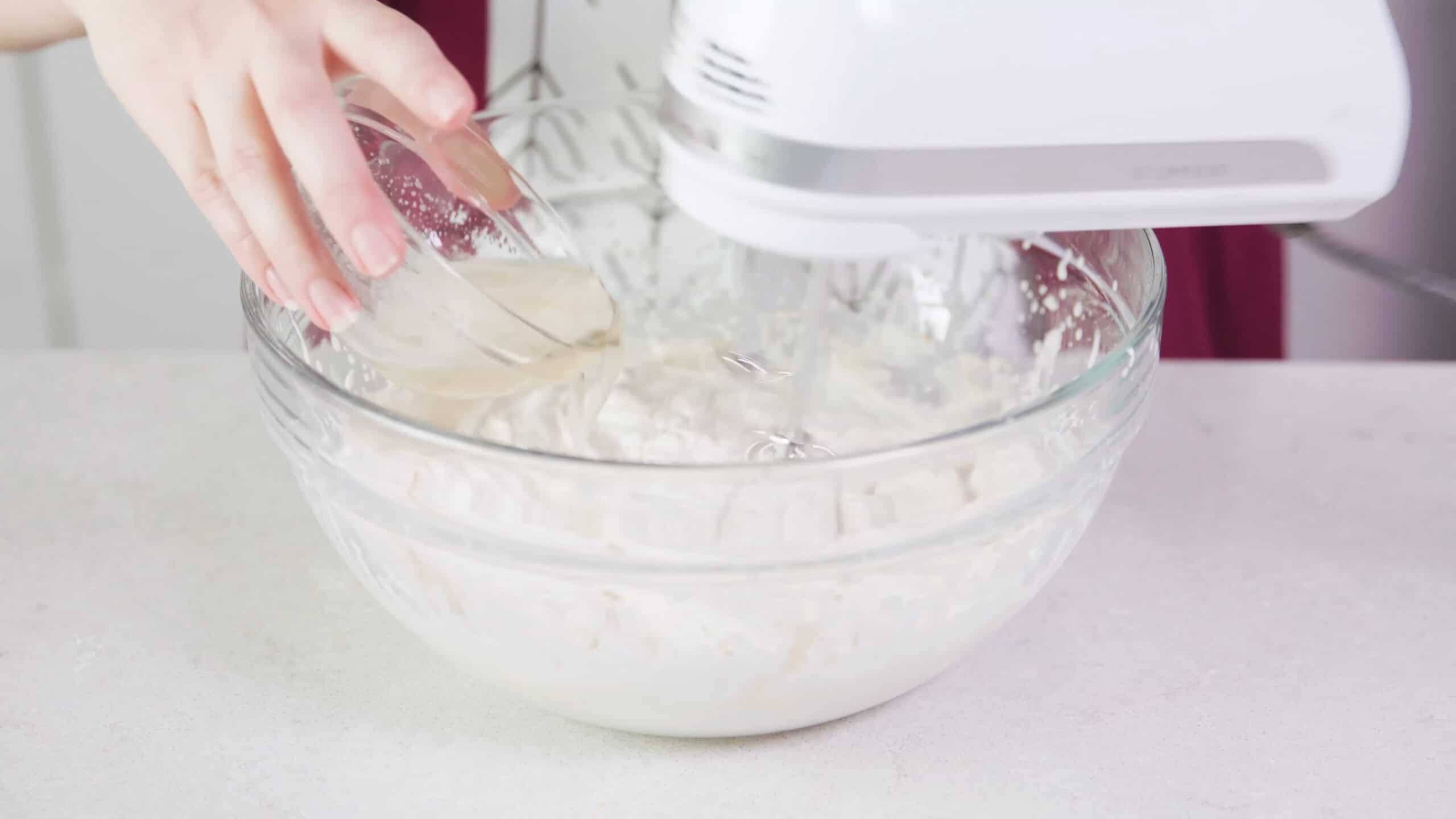Angled view of large glass mixing bowl filled with whipped cream while pouring from a small glass bowl filled with gelatin being mixed with a hand mixer to stabilize the whipping cream.