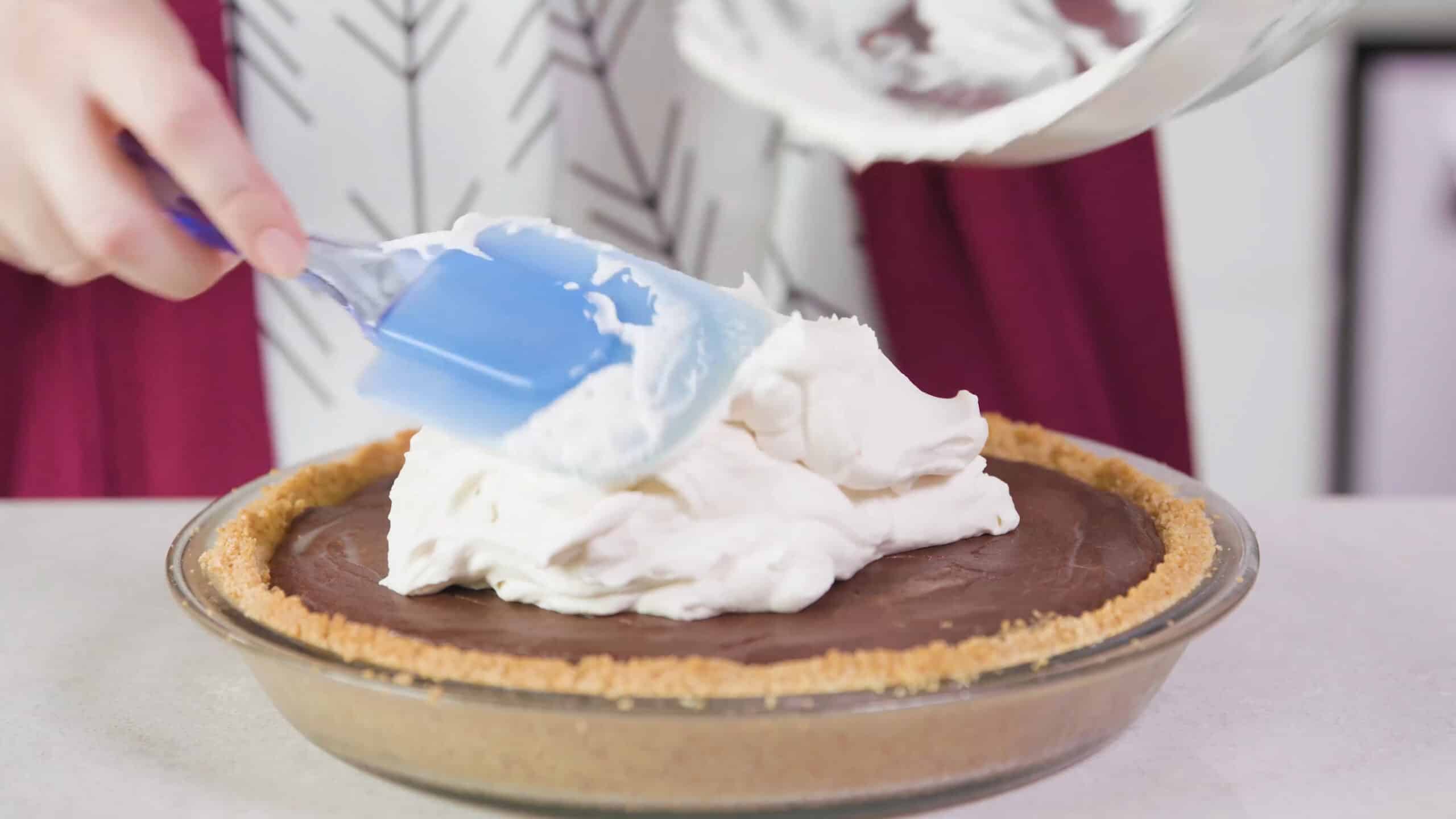 Side view of cooled chocolate cream pie with whipped cream piled on over the top by a plastic spatula from a clear glass mixing bowl.
